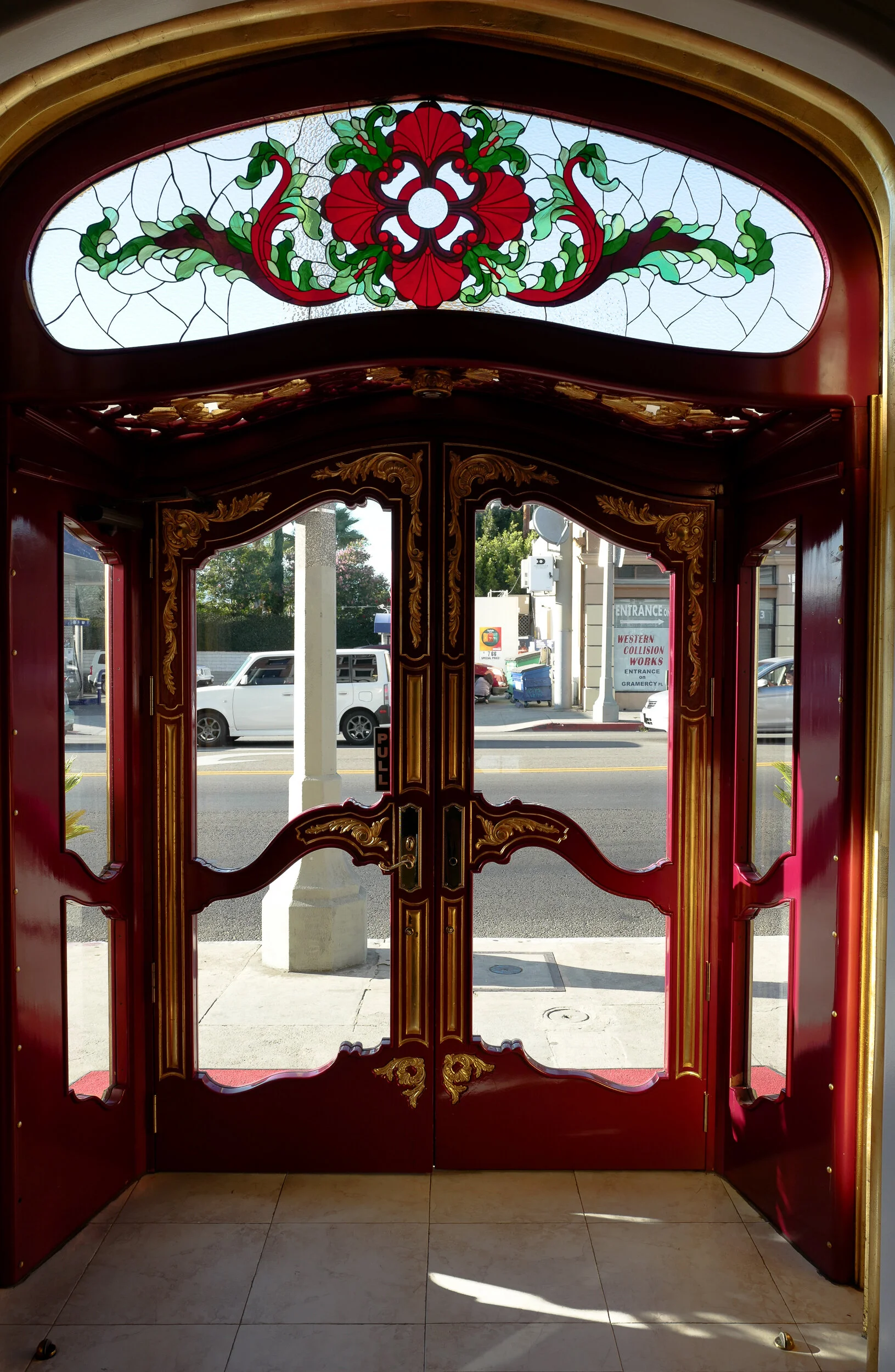 Decorative red double doors with ornate gold accents and stained glass with a floral design at the top, leading outside to a street view with parked cars and buildings.