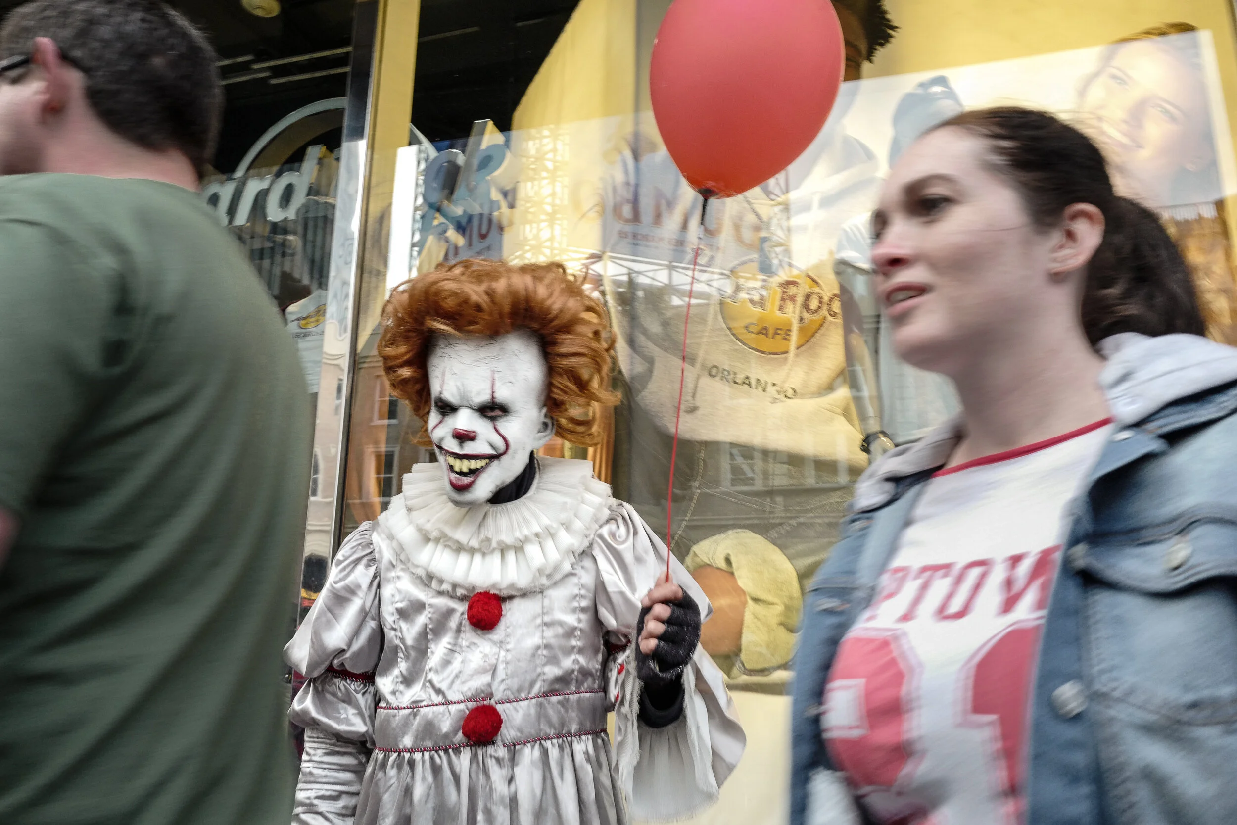 A person dressed as Pennywise the clown from IT, with white face paint, red hair, and a ruffled costume, holding a red balloon, standing on a busy street with pedestrians passing by.