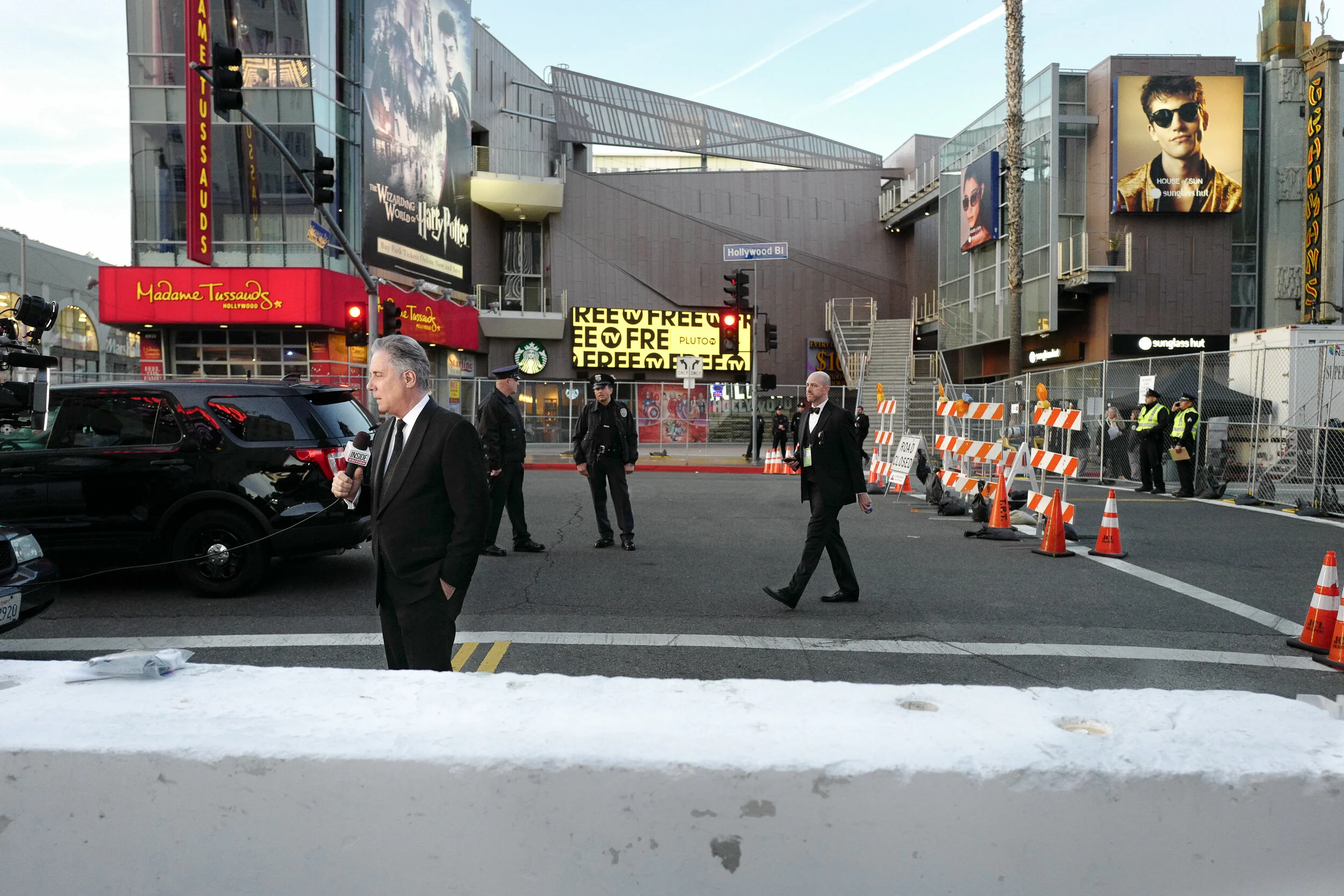 Street scene at night with a man holding a microphone, police officers, barricades, and police tape, set in an area with bright neon signs and billboards, including Madame Tussauds and Harry Potter.