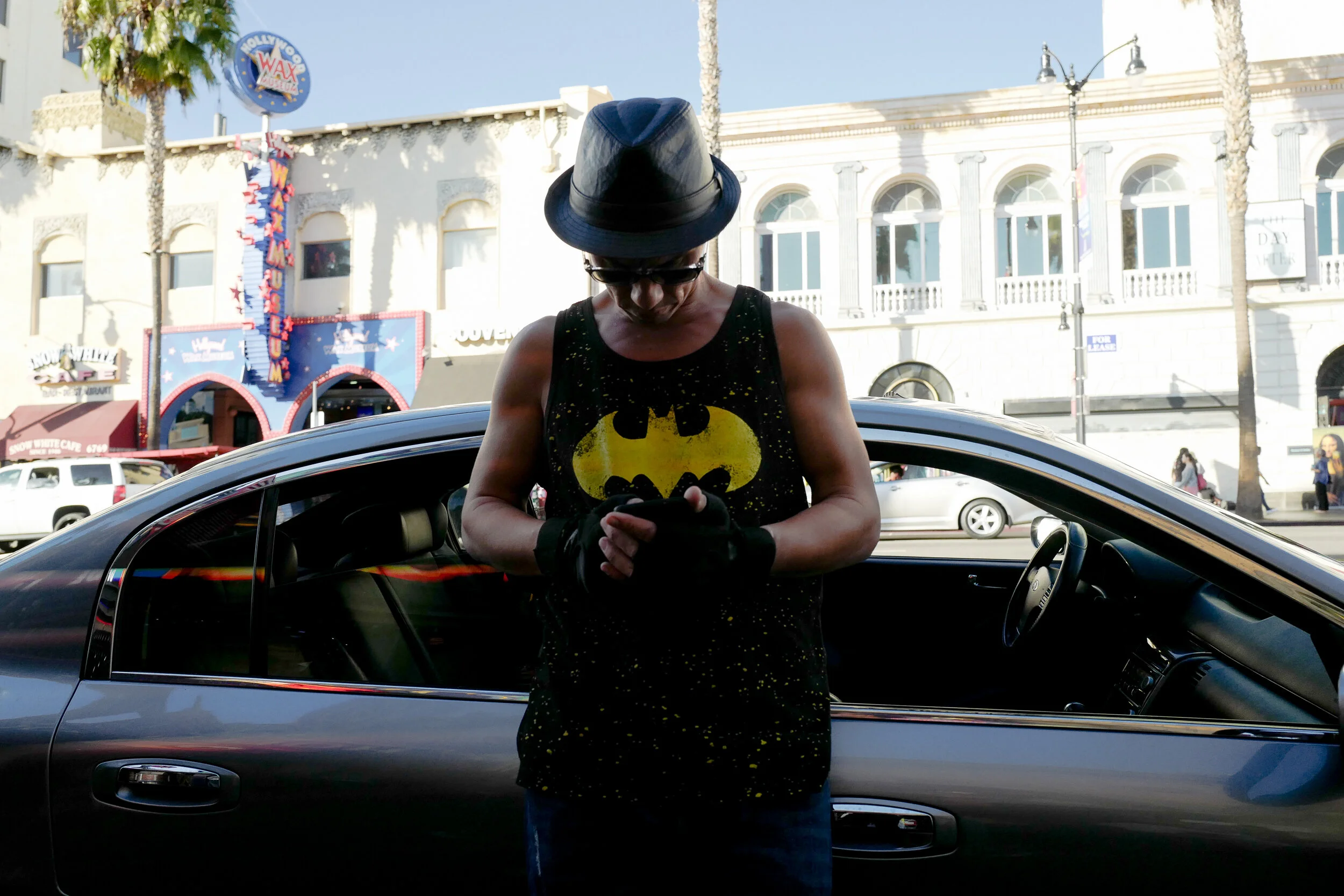A person wearing a Batman tank top, black gloves, and a fedora hat stands beside a black car, looking at their phone, with a street and buildings in the background.