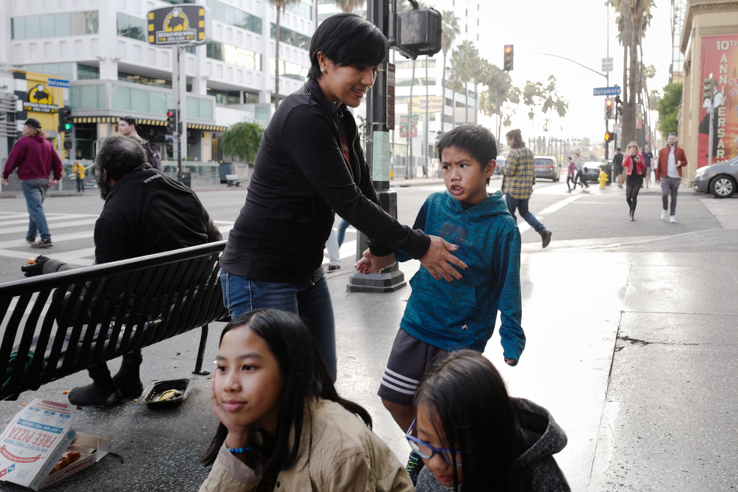 A woman is helping an upset boy on a city street while three girls sit nearby. Pedestrians pass by, and buildings line the street.