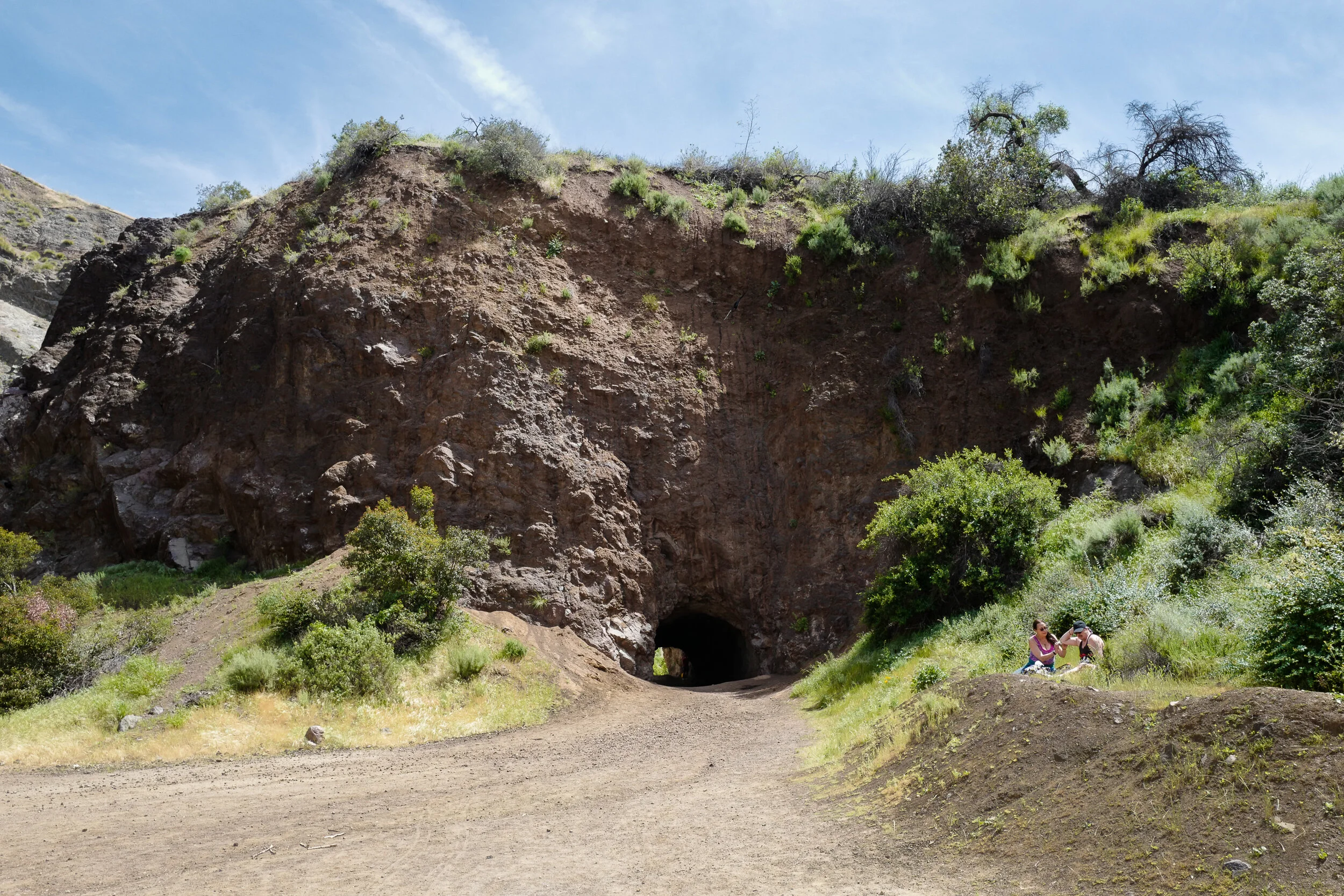 Two women sitting on a grassy mound near a trail, with a large rocky hill and tunnel in the background, surrounded by green bushes and trees.