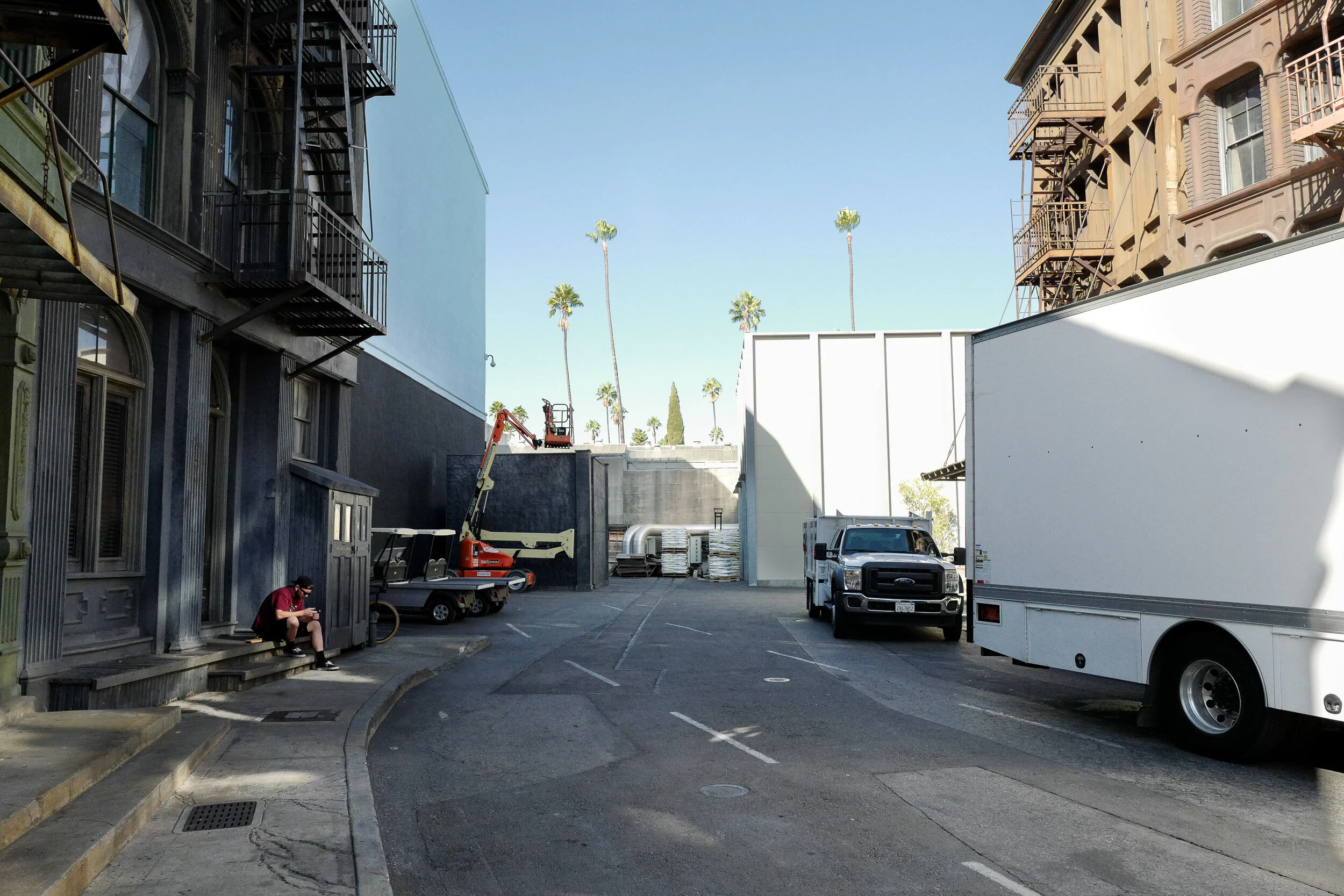 A narrow city alley with parked trucks, construction equipment, and a man sitting on a step using his phone. Tall palm trees are visible in the background against a clear blue sky.