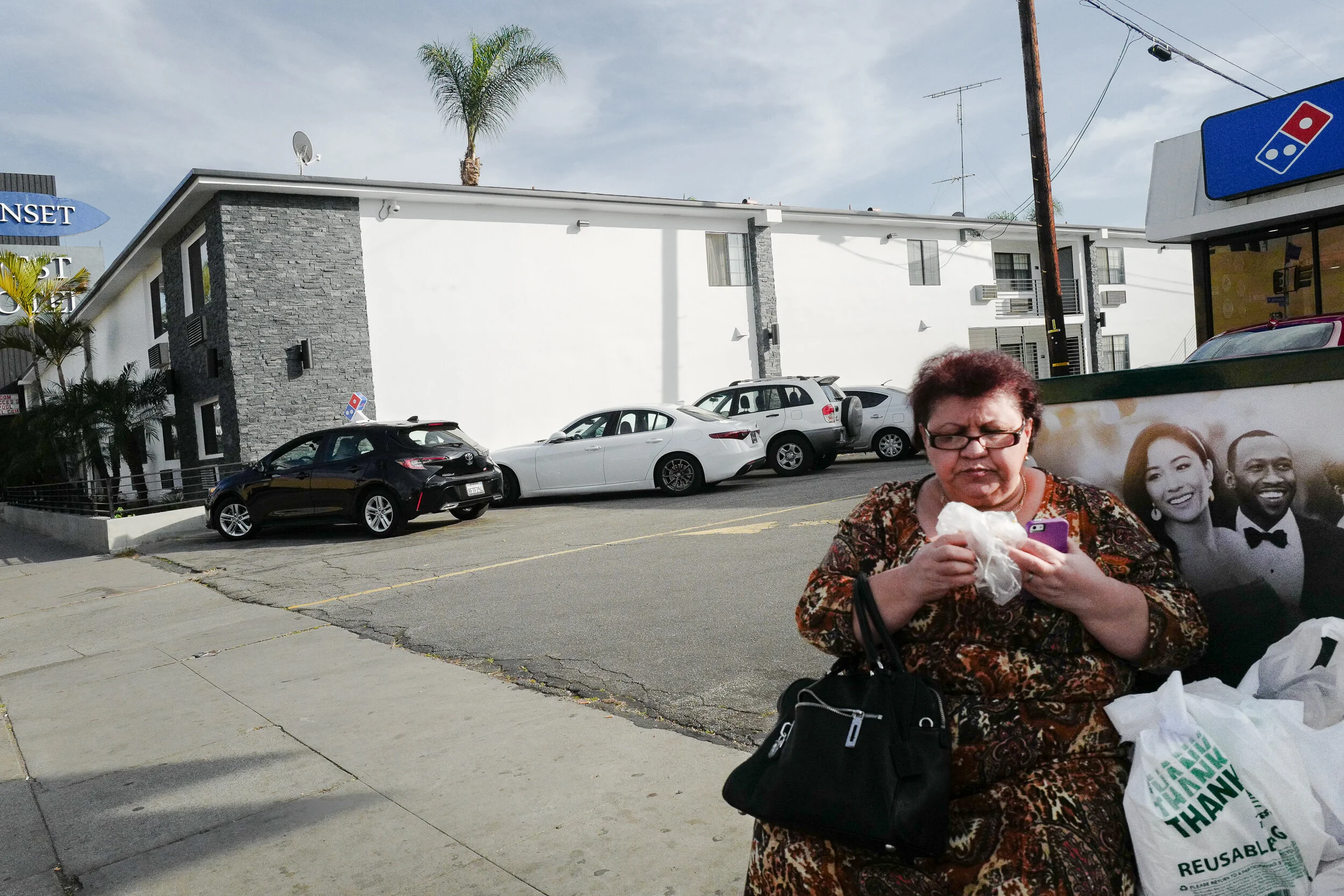 A woman sitting on a bench outside a pizza restaurant, looking at her phone and holding a plastic bag, with parked cars and a white building in the background.