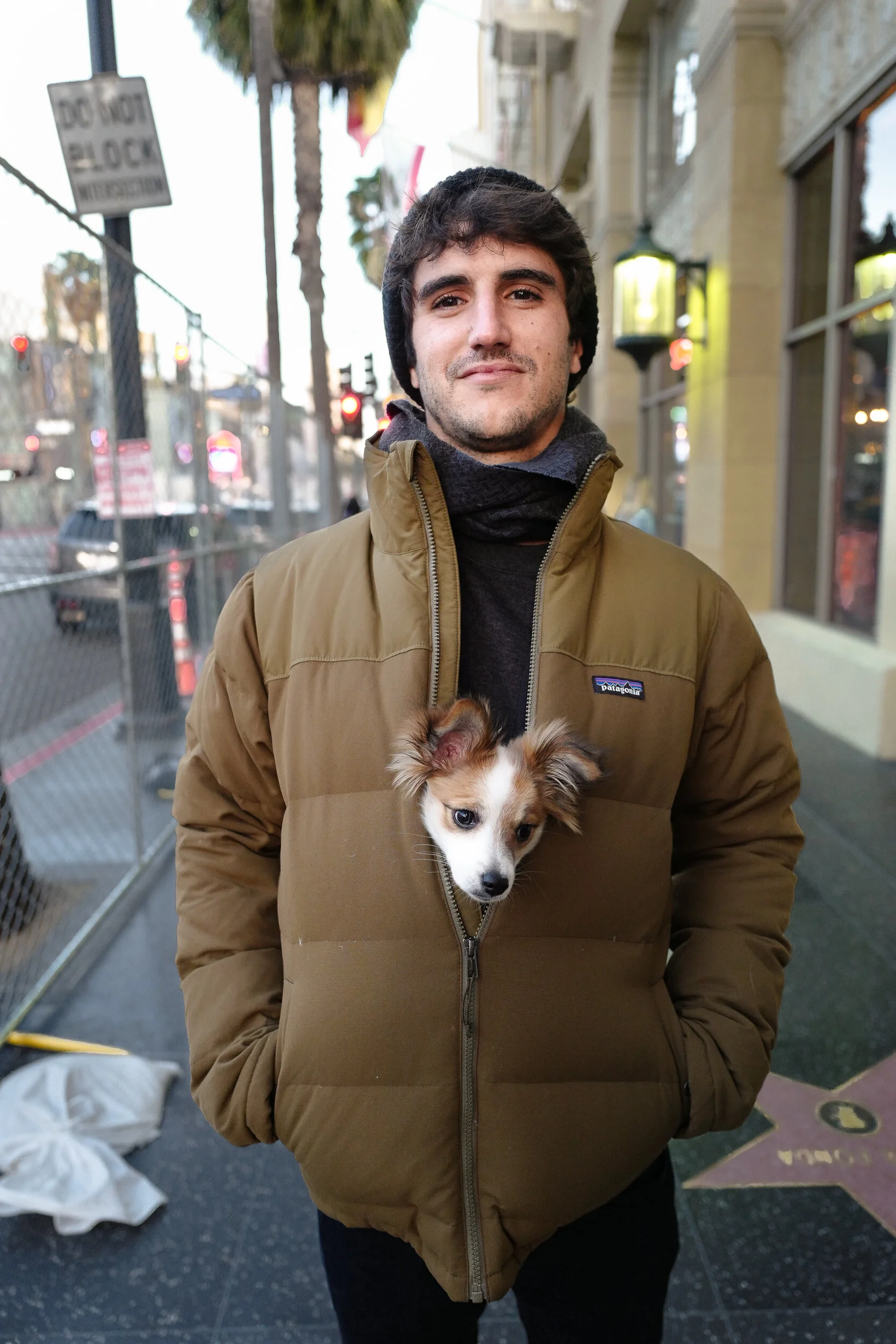 A young man with dark hair and a slight beard standing outdoors on a city street, wearing a brown jacket with a puppy peeking out from the unzipped front pocket.