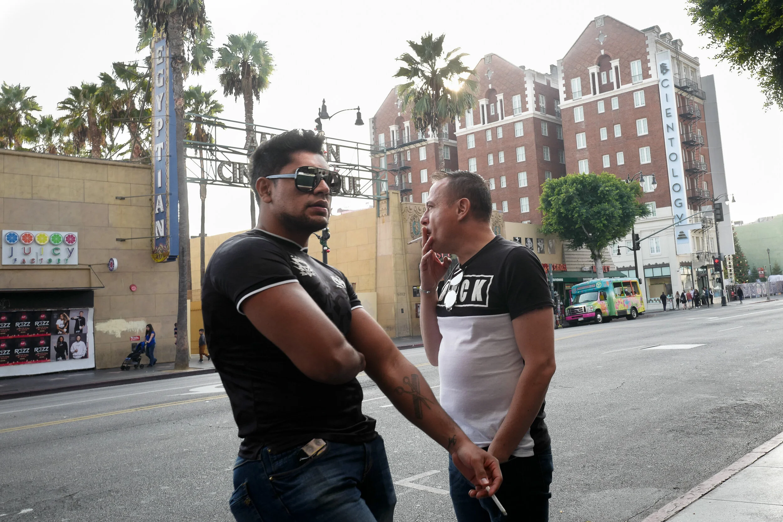 Two men standing on a city street in front of the Magic Johnson Theater, one wearing sunglasses and a black shirt, the other smoking and talking, with palm trees and a colorful van in the background.
