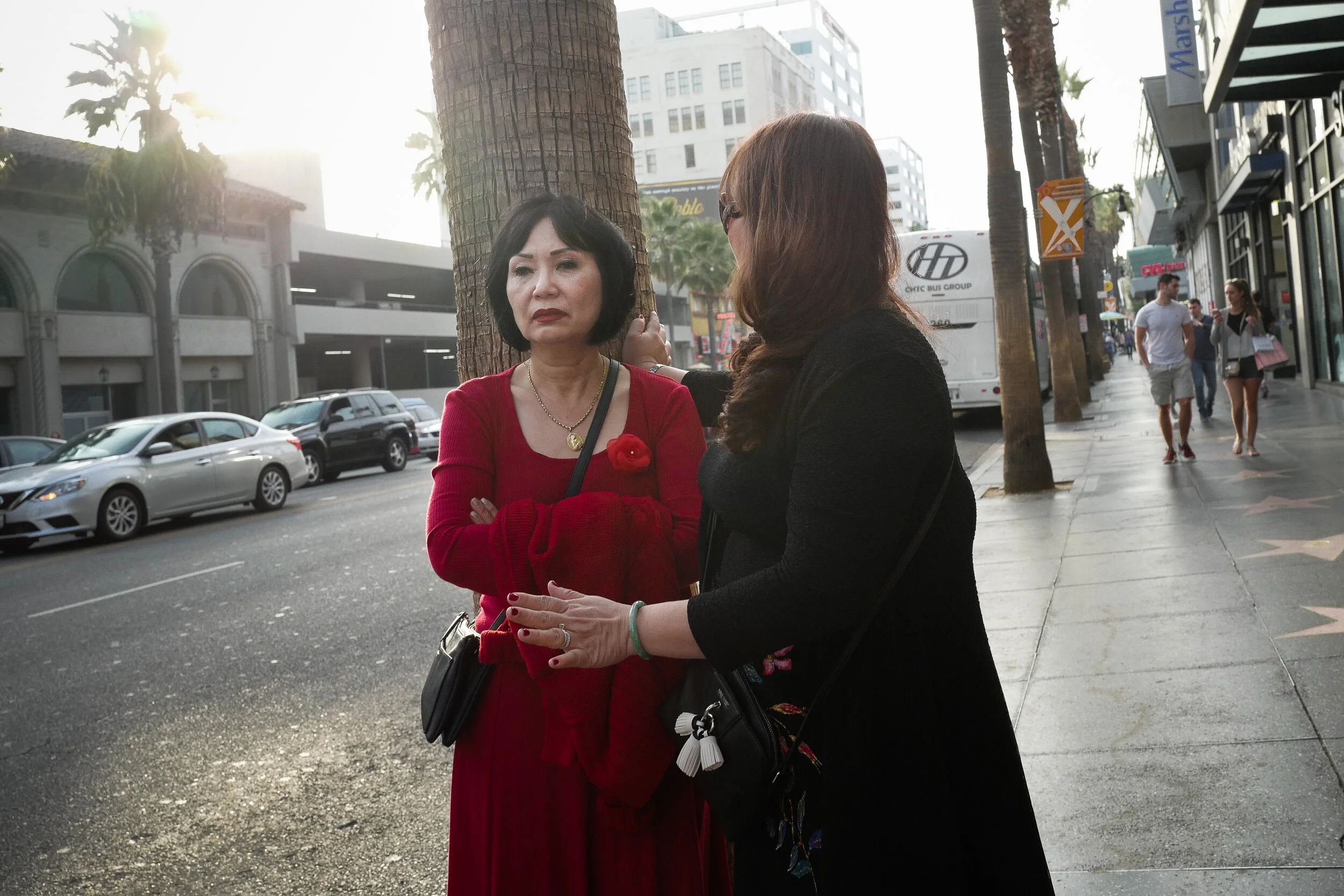 Two women having a serious conversation on a city sidewalk, with palm trees, cars, and pedestrians in the background.