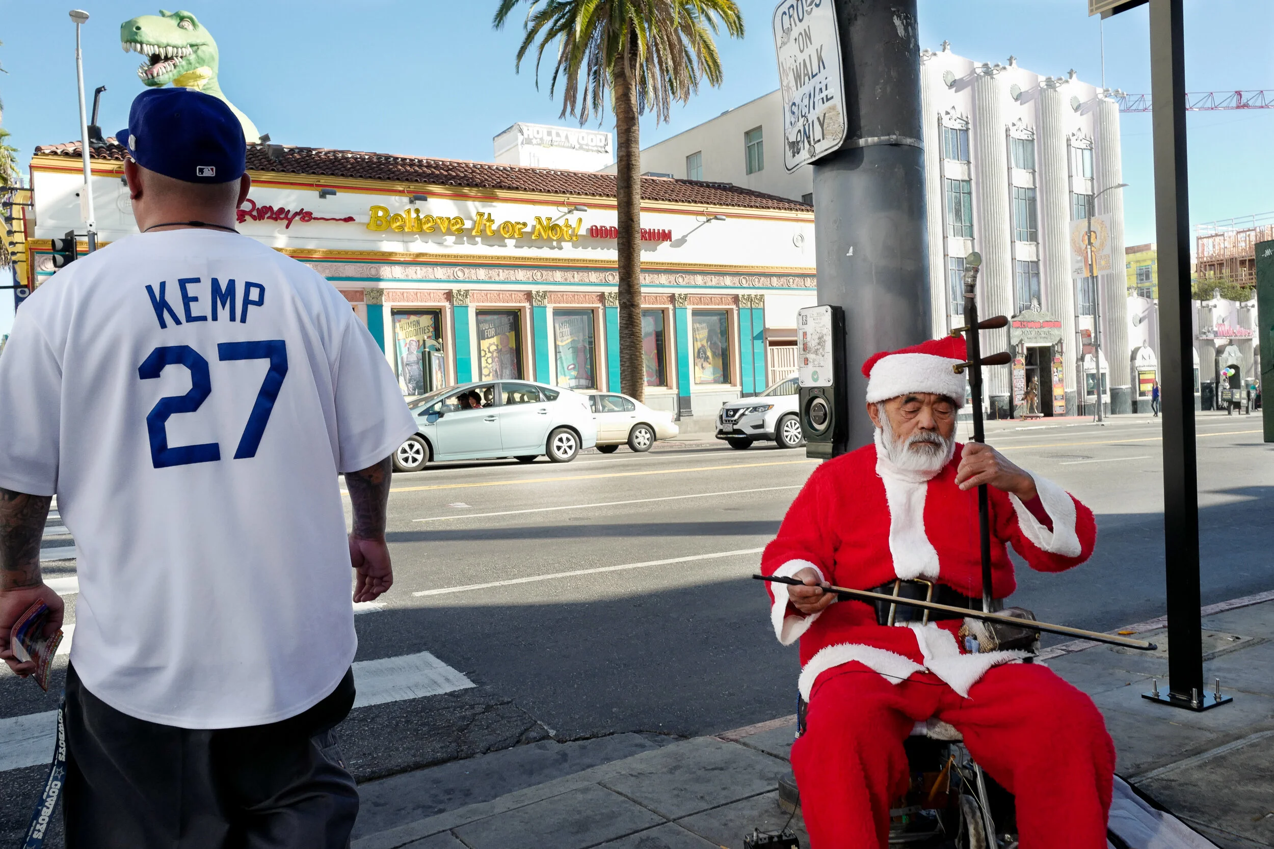 A man dressed as Santa Claus sitting on the sidewalk playing a musical instrument, while a person in a white sports jersey and blue cap walks past on the street. Buildings, cars, and a palm tree are visible in the background.