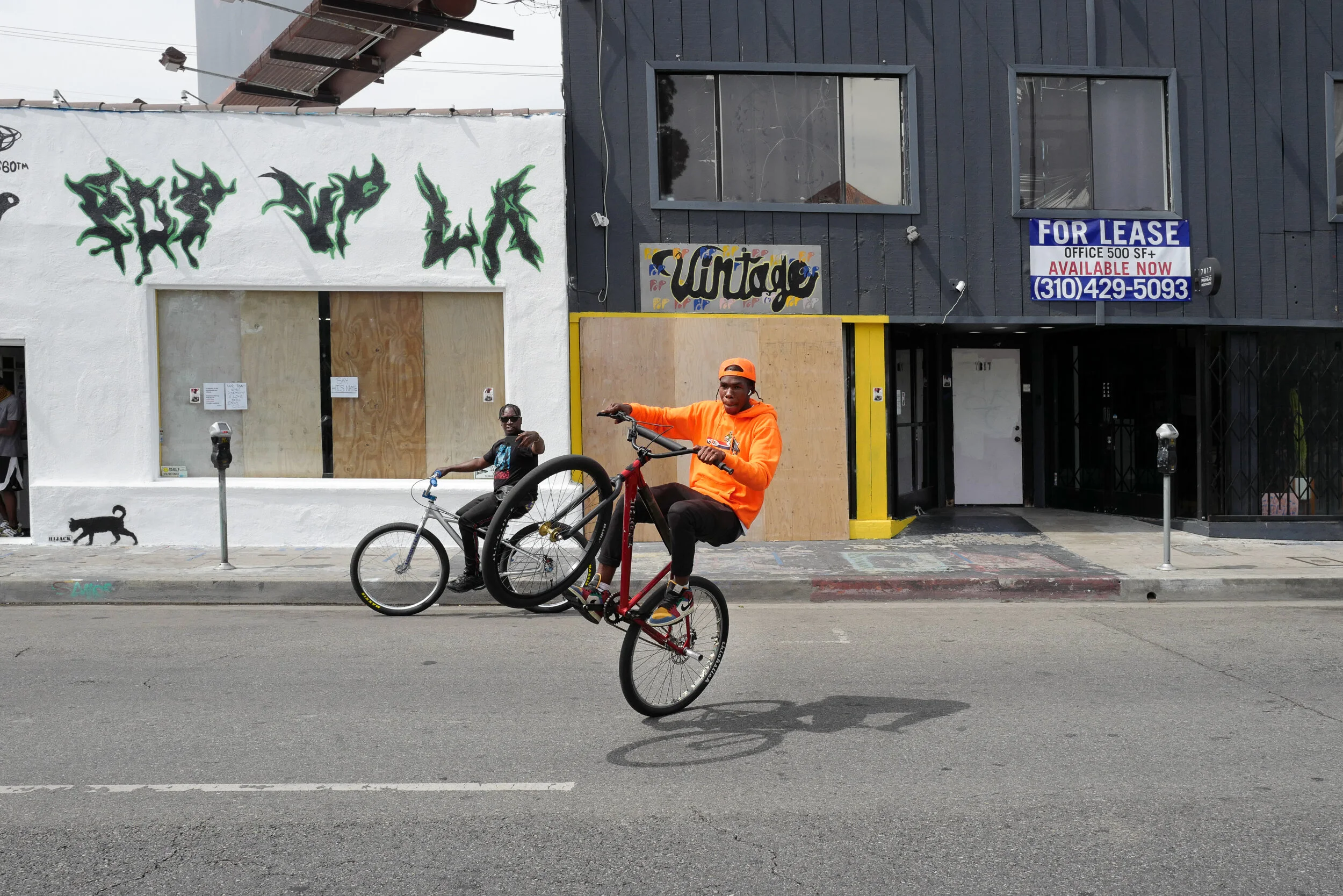 A man performing a wheelie on a red BMX bike on the street, wearing an orange hoodie and cap. In the background, a person is sitting on a bicycle, and there are buildings with a 'For Lease' sign, graffiti, and a parking meter.