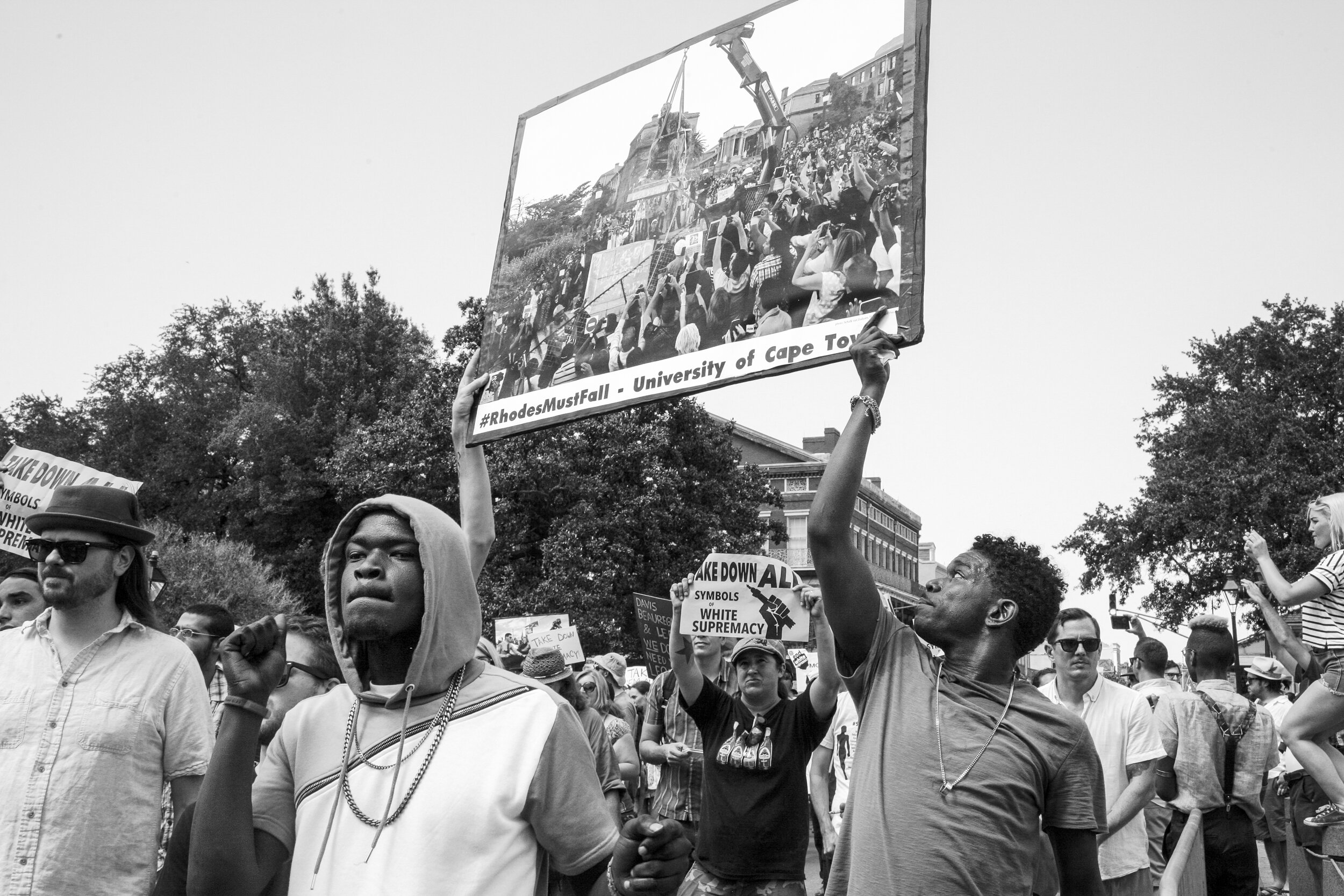 Protestors participating in march, holding a sign drawing connection to the removal of Apartheid monuments in South Africa 