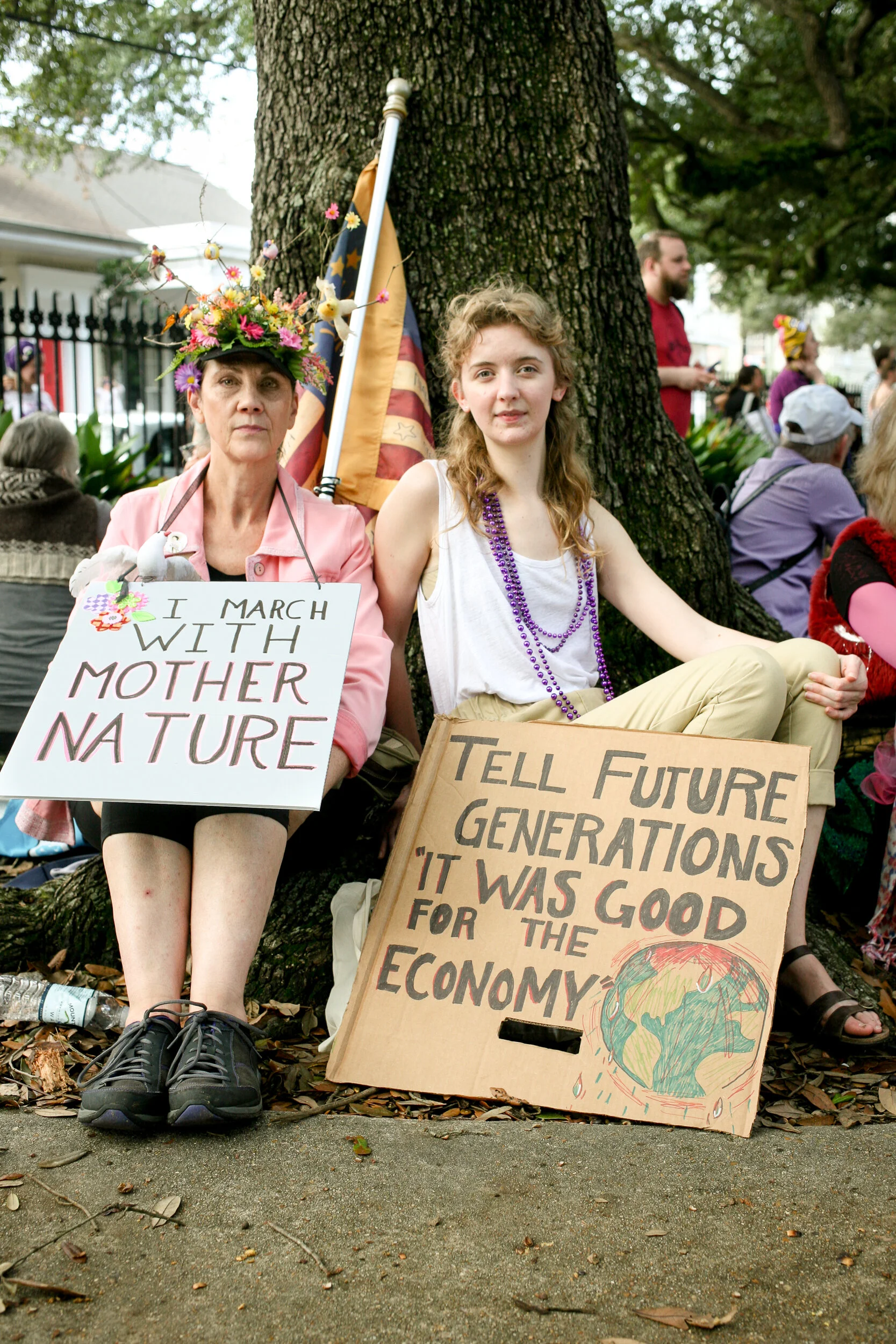 Two women sitting under a tree at a protest. One woman wears a flower crown and holds a sign that says 'I March With Mother Nature.' The other woman holds a cardboard sign that reads 'Tell Future Generations 'It Was Good For The Economy' with a drawi