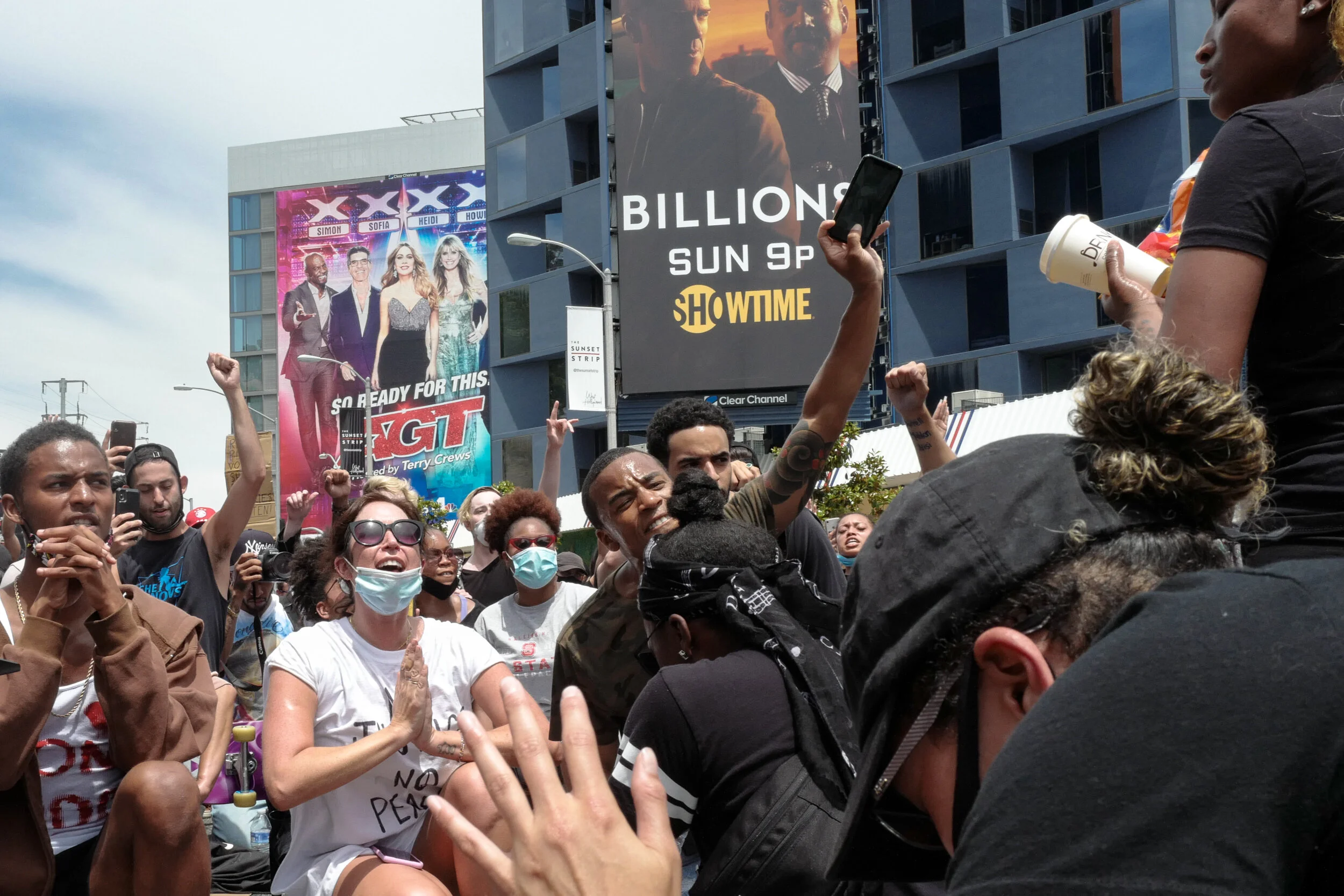 A diverse group of people protesting or demonstrating outdoors, some with raised fists, wearing masks, and holding signs, with billboards and a building in the background.