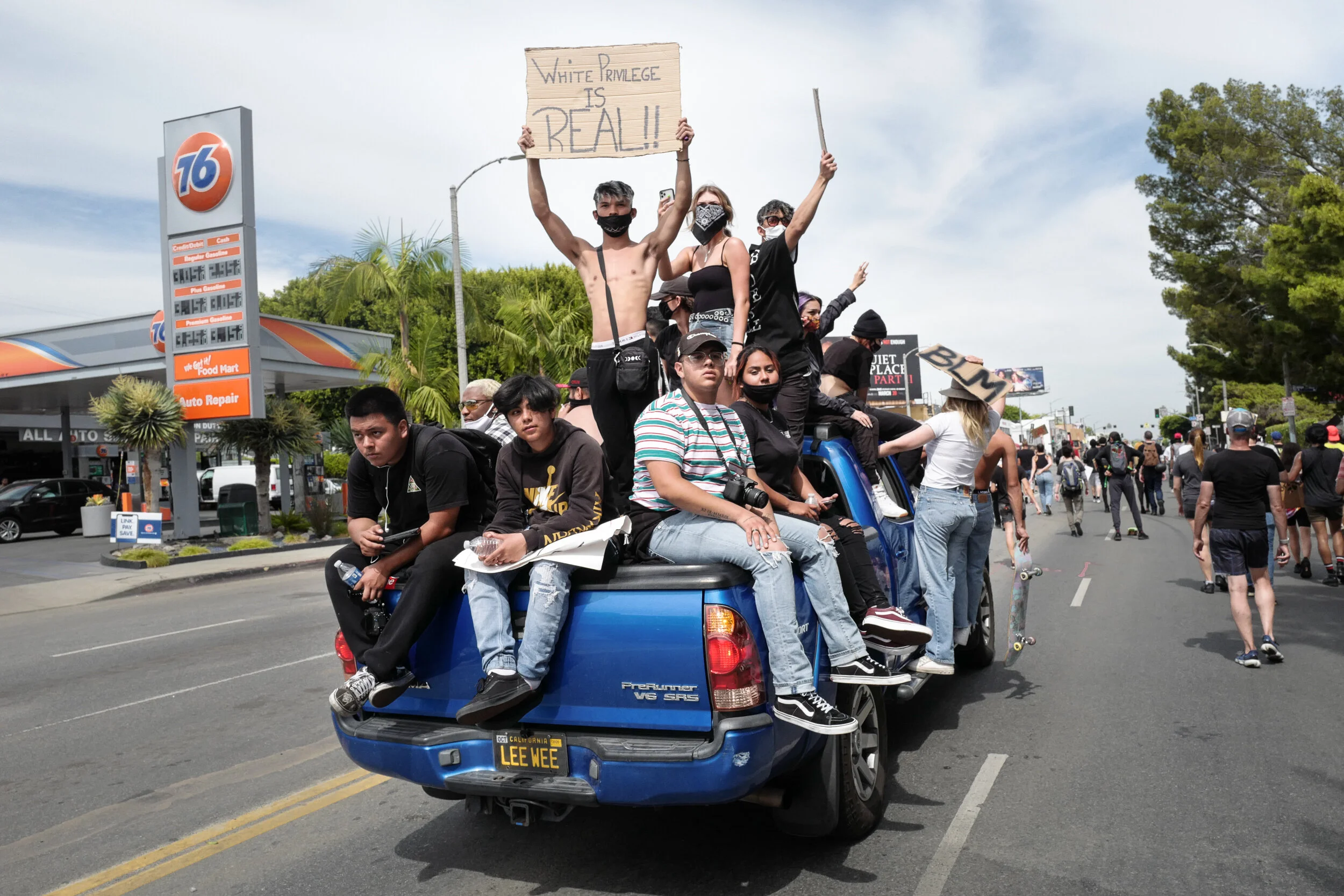 Protesters sitting and standing on a blue pickup truck in a street protest. One person holds a sign stating 'White Privilege is Real!!' Others are holding signs or skateboarding.