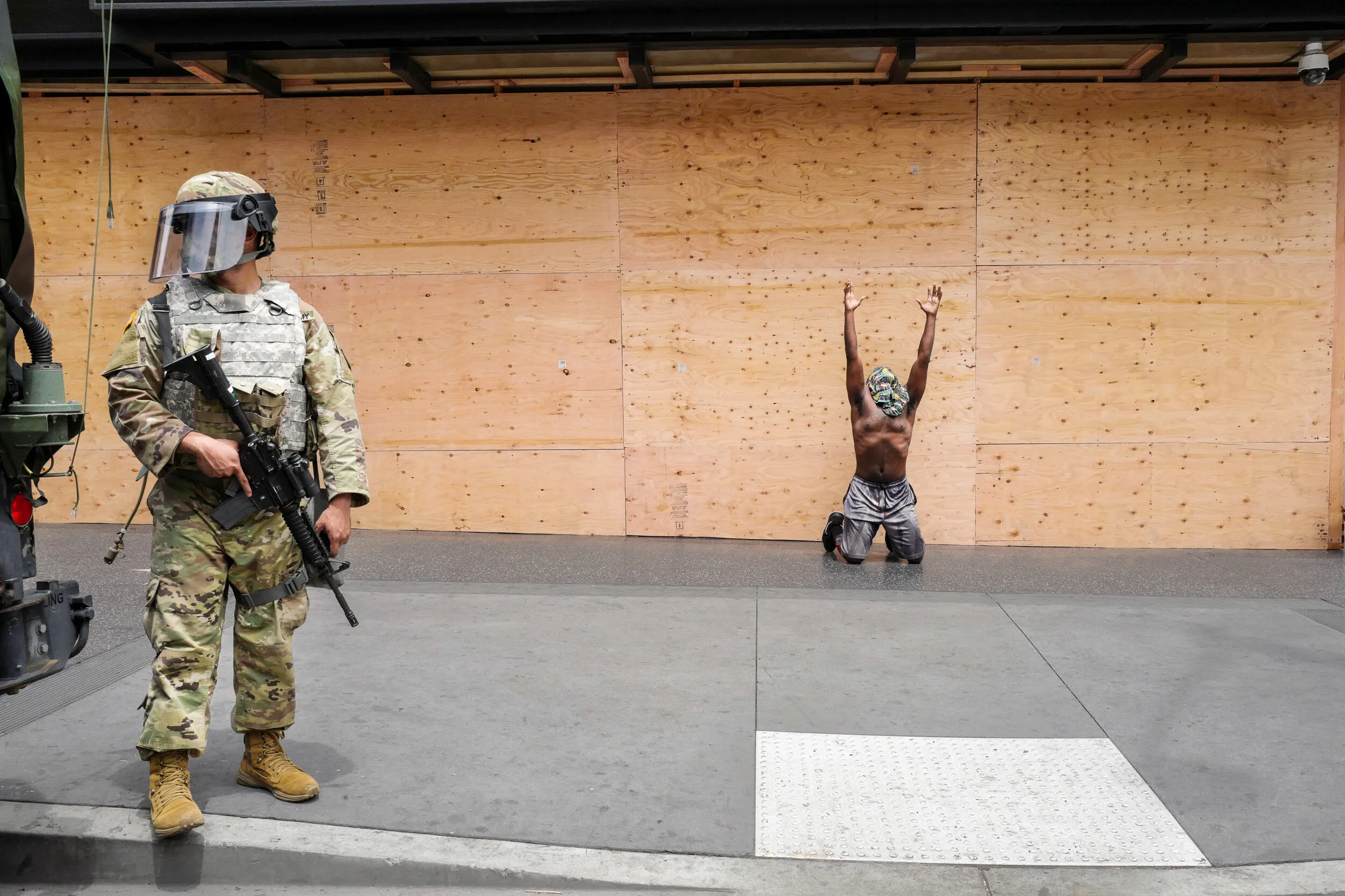 A soldier in camouflage uniform and riot gear stands at attention with a rifle, while a man kneels on the ground with his arms raised, wearing a colorful mask and gray shorts, in front of a plain wooden wall.