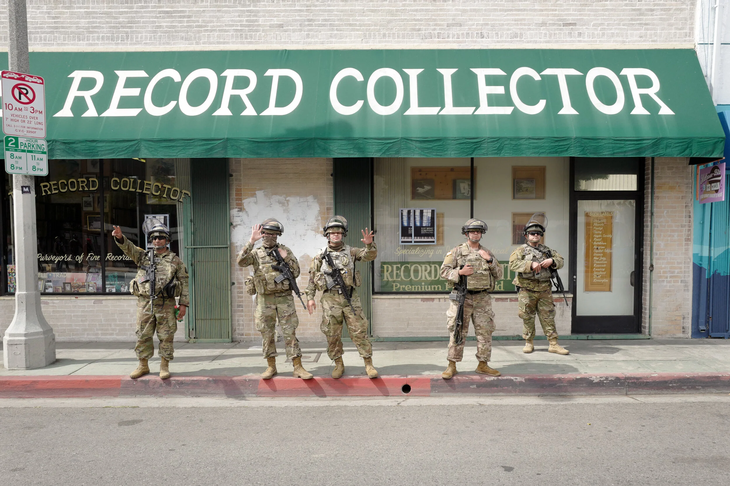 Five soldiers in camouflage uniforms and tactical gear standing in front of a record store with a green awning that reads 'Record Collector'. The soldiers are wearing helmets, sunglasses, and carrying rifles, with some waving and others with weapons 
