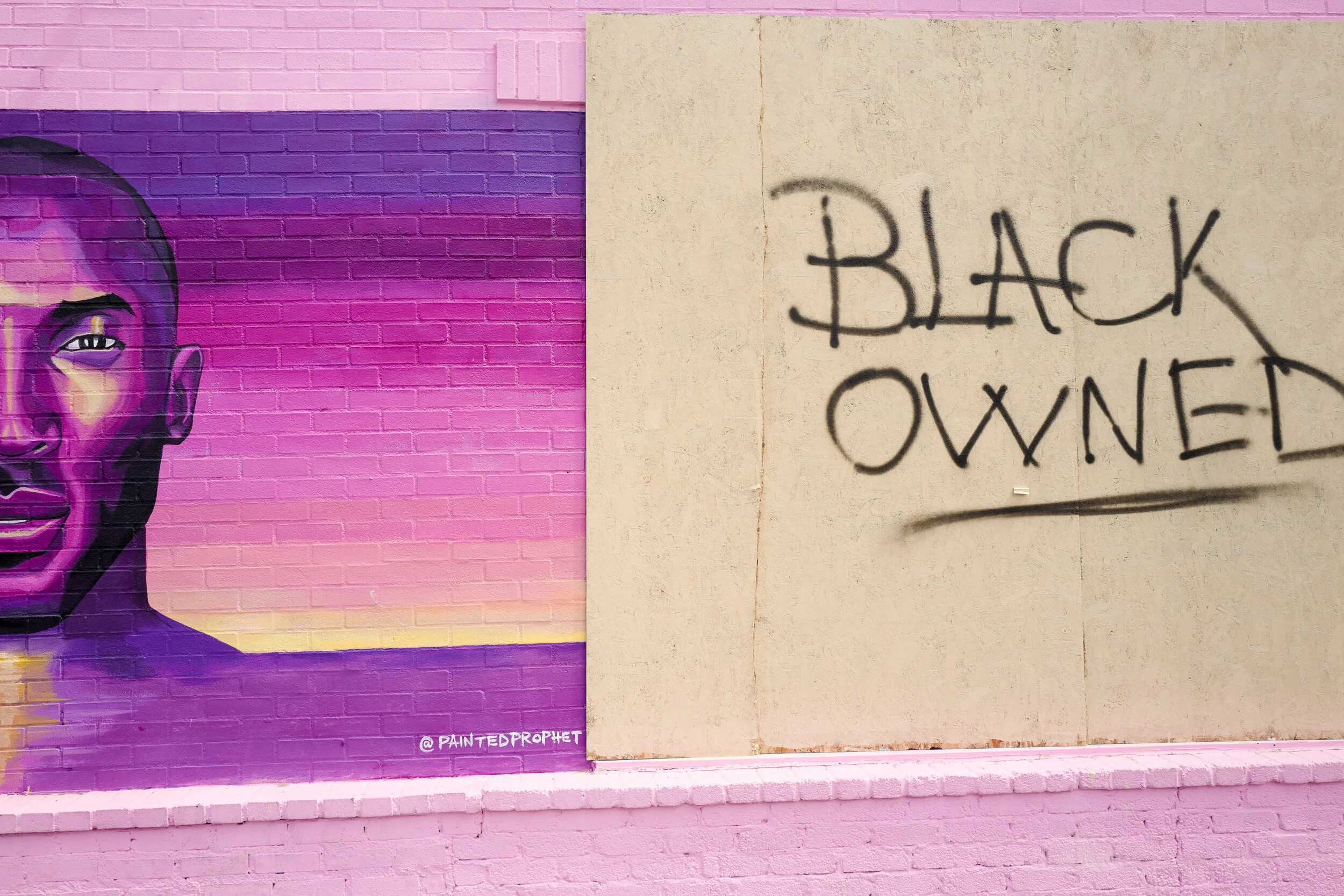 A mural of a person's portrait painted on a brick wall with pink, purple, and yellow background, next to a beige wall with black graffiti that reads 'Black Owned'.