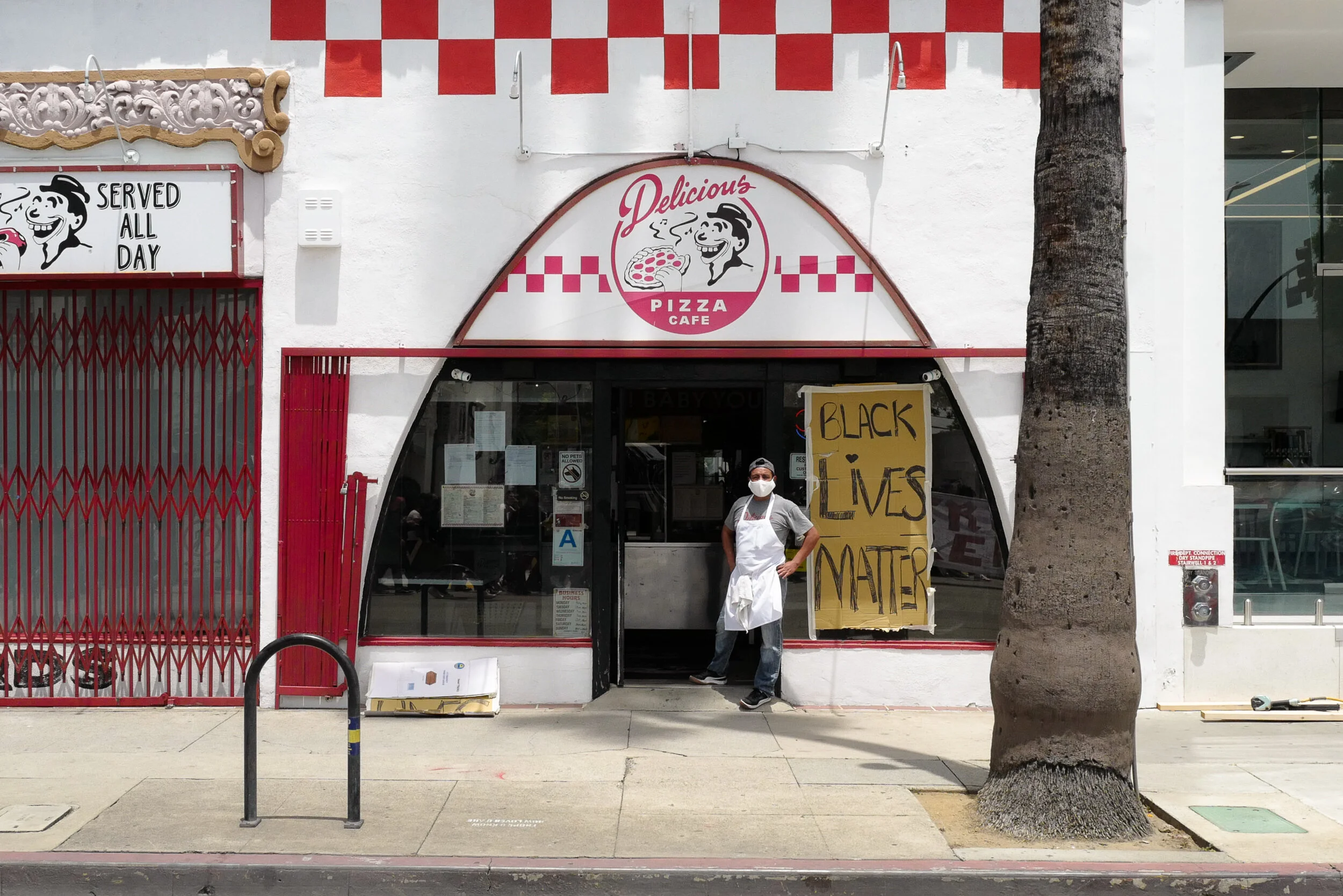 A man standing outside a pizza cafe with a sign that says 'Black Lives Matter' on the front window. The cafe has a red and white checkered sign that says 'Delicious Pizza Cafe'. The man is wearing a mask, apron, gray shirt, and jeans, standing next t