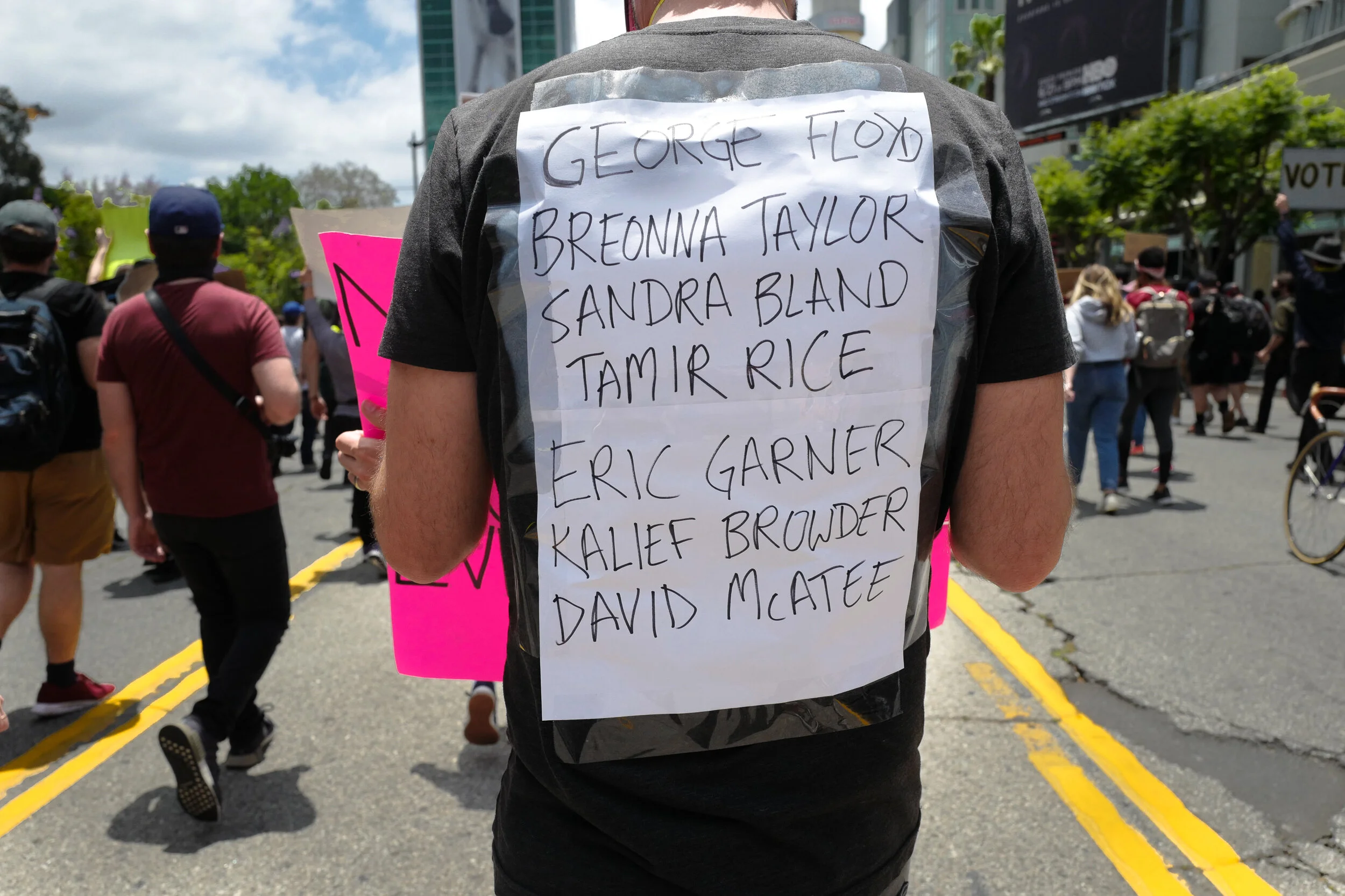 A person protesting at a rally, wearing a vest with a sign listing names of victims. The background shows other protesters holding signs on a city street.