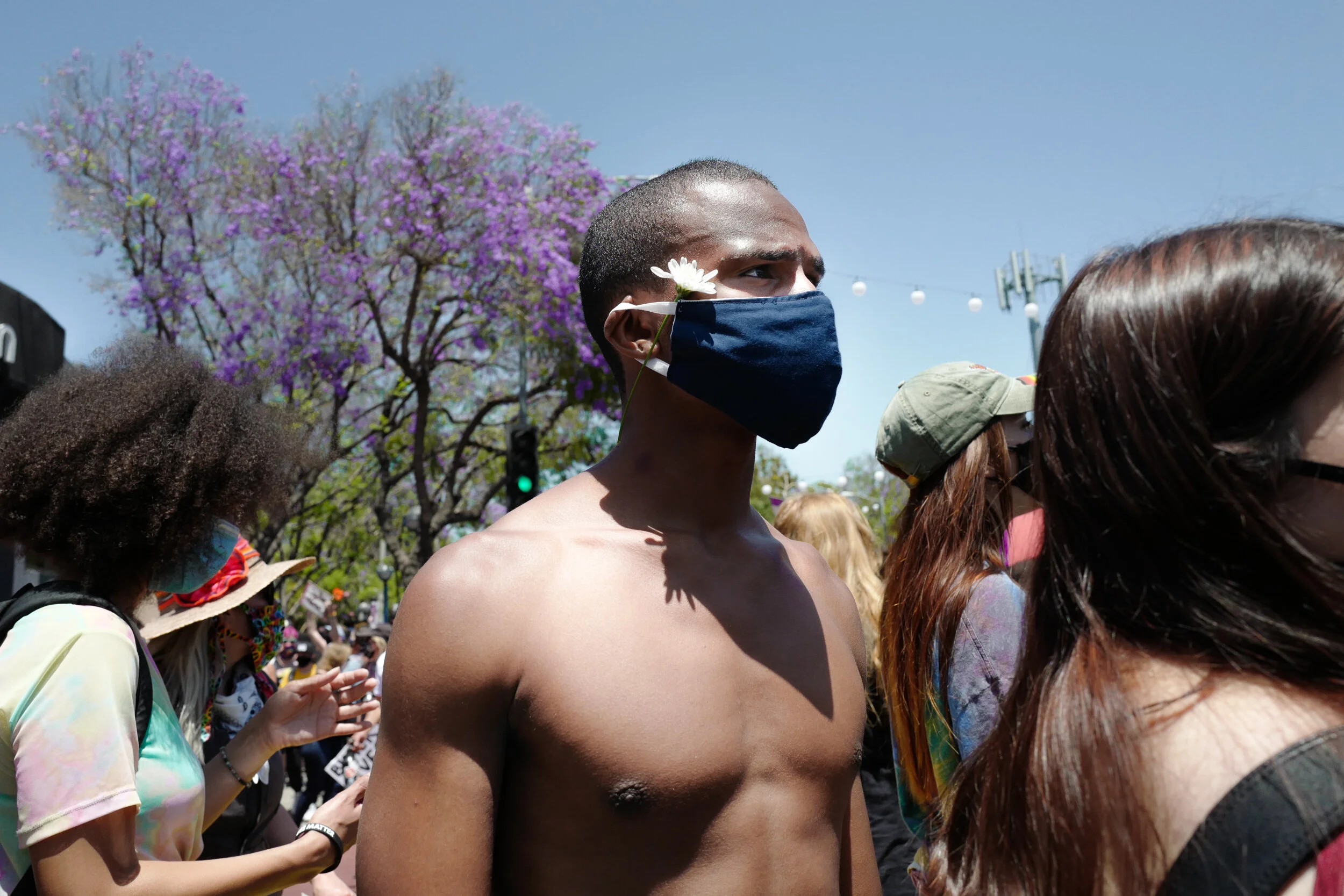 A shirtless person wearing a black face mask with a white flower attached, participating in an outdoor gathering or protest, with other people around, some wearing masks, and a large purple-flowered tree in the background.