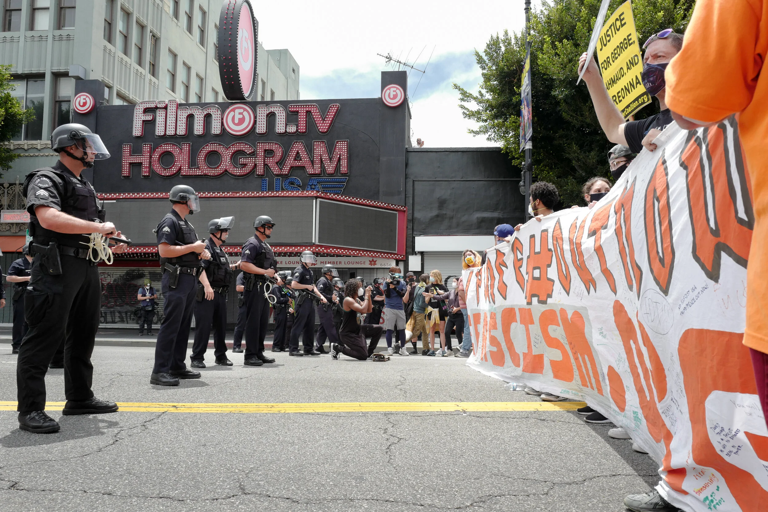 A group of police officers in riot gear facing a line of protesters holding a large banner with messages. The scene appears to be part of a demonstration outside a building with a sign reading 'Fremont TV Hologram'.