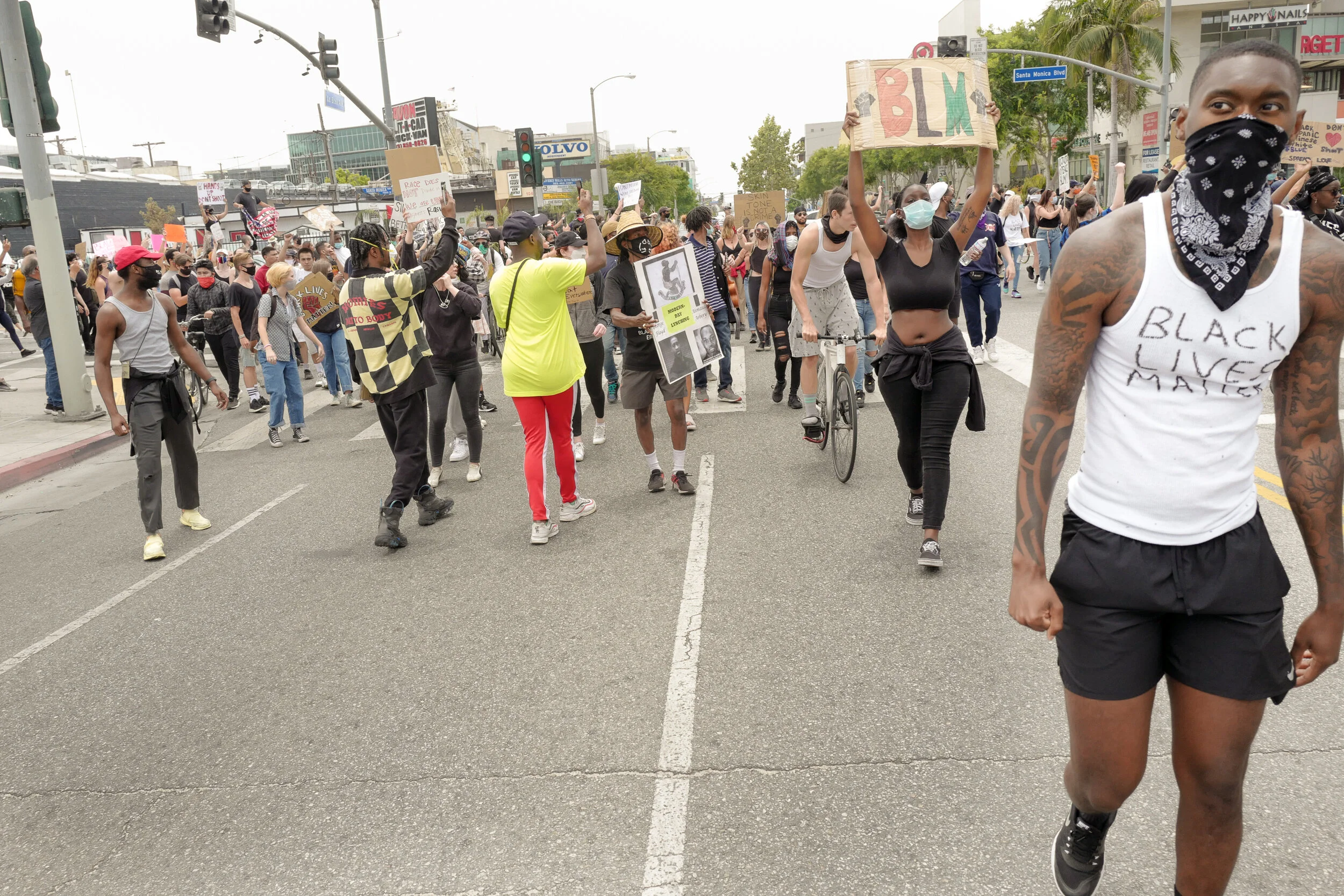 A large group of protesters marching on a city street during a demonstration, many holding signs including one with the letters 'BLM' and wearing masks, with some wearing shirts with messages supporting Black Lives Matter.
