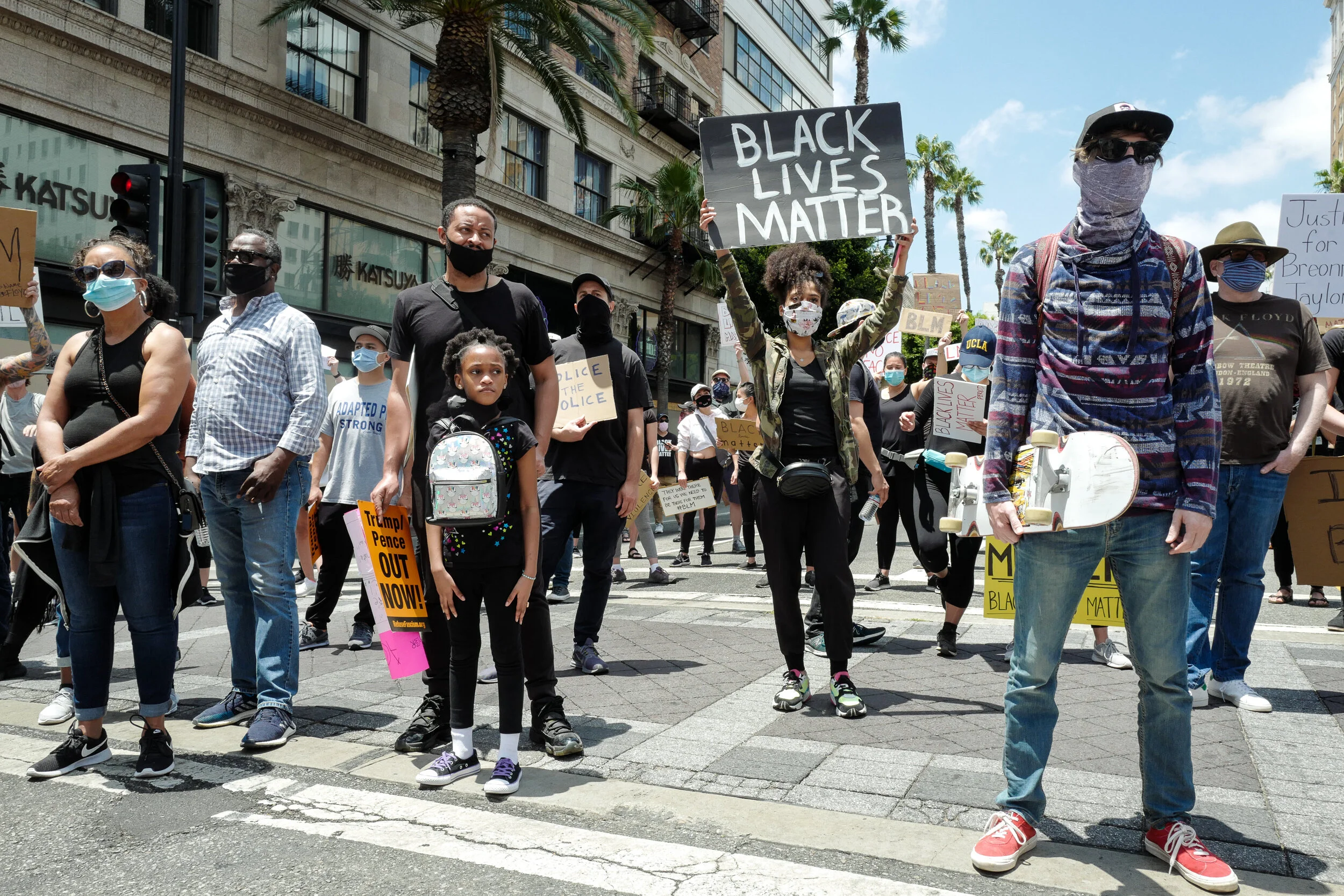 A diverse group of people protesting in the street, many holding signs with messages like "Black Lives Matter." Some people are wearing masks and sunglasses, with a man holding a skateboard in the foreground and a woman holding a "Black Lives Matter"