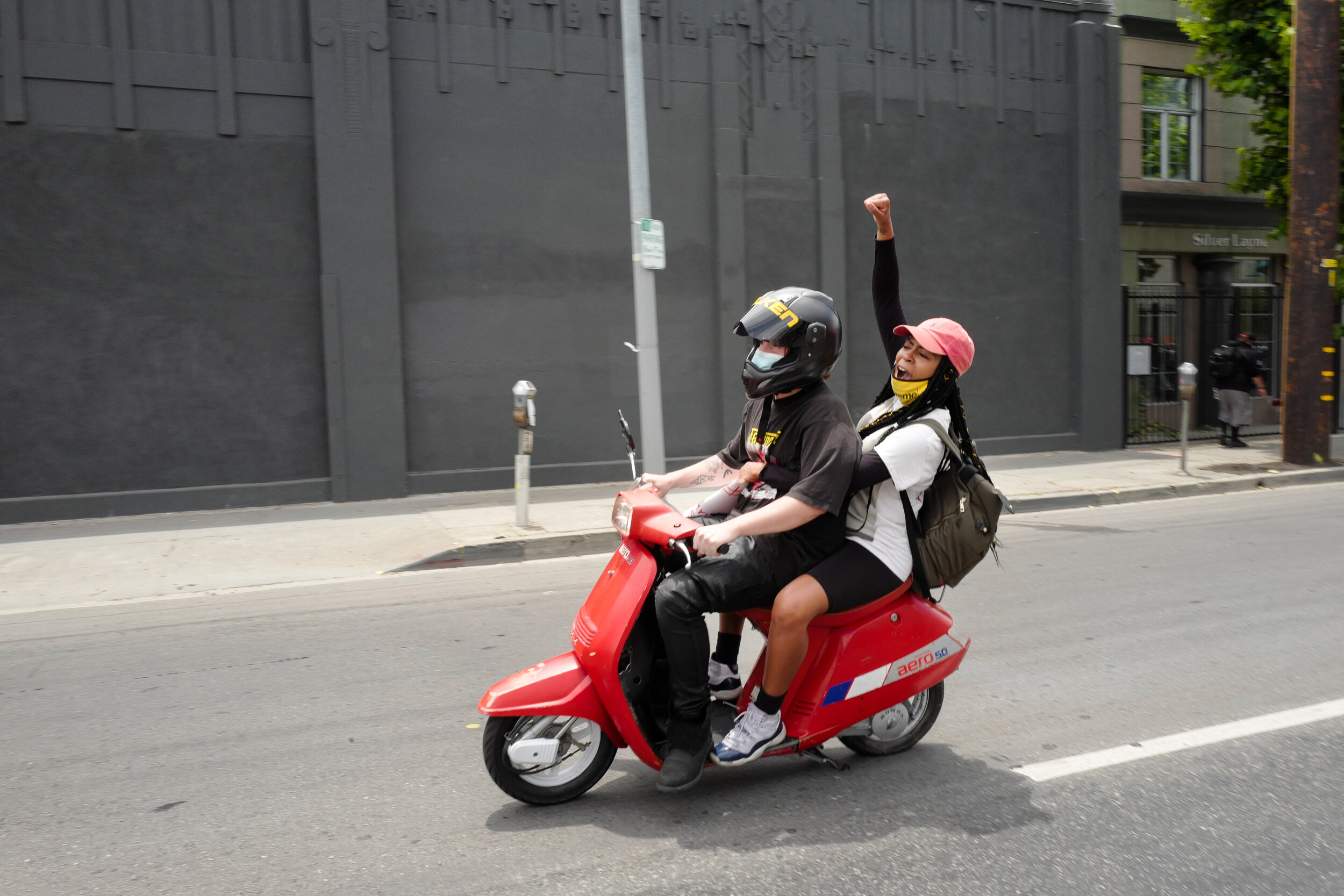 A person driving a red scooter with a passenger, both wearing helmets, on a city street. The passenger is raising an arm and appears to be celebrating.