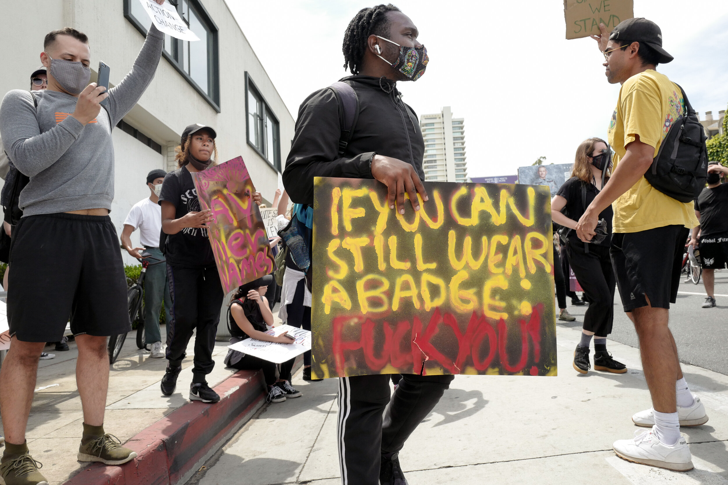 Protesters gathered outdoors, some holding signs, including a person in the center holding a spray-painted cardboard sign that reads, "IF YOU CAN STILL WEAR A BADGE: FUCK YOU." Others are taking photos or engaged in conversation, with buildings in th