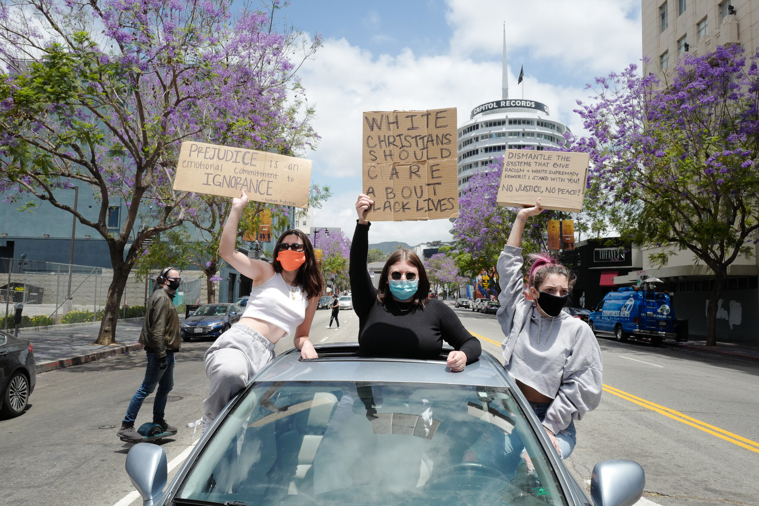 Three women standing through the sunroof of a car protesting on a city street with purple trees in the background. They hold signs with messages about racial justice, including one that reads 'WHITE CHRISTIANS SHOULD CARE ABOUT BLACK LIVES.' All wome