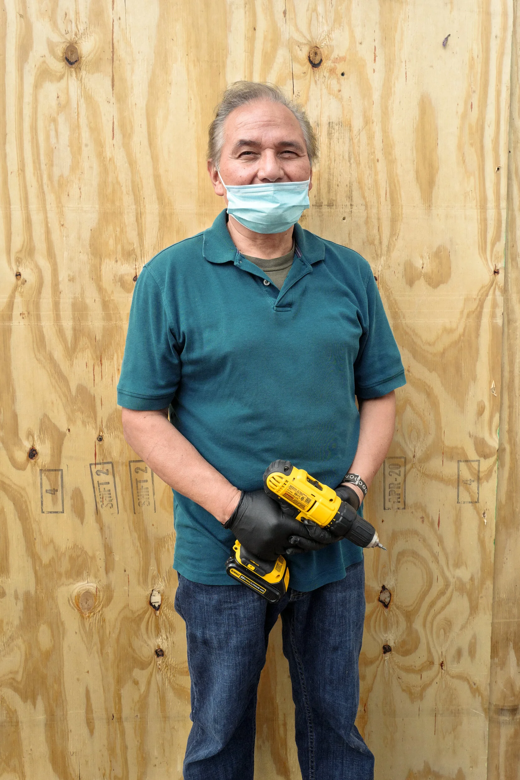 Man wearing a face mask and gloves, holding a yellow cordless drill, standing in front of a wooden wall.