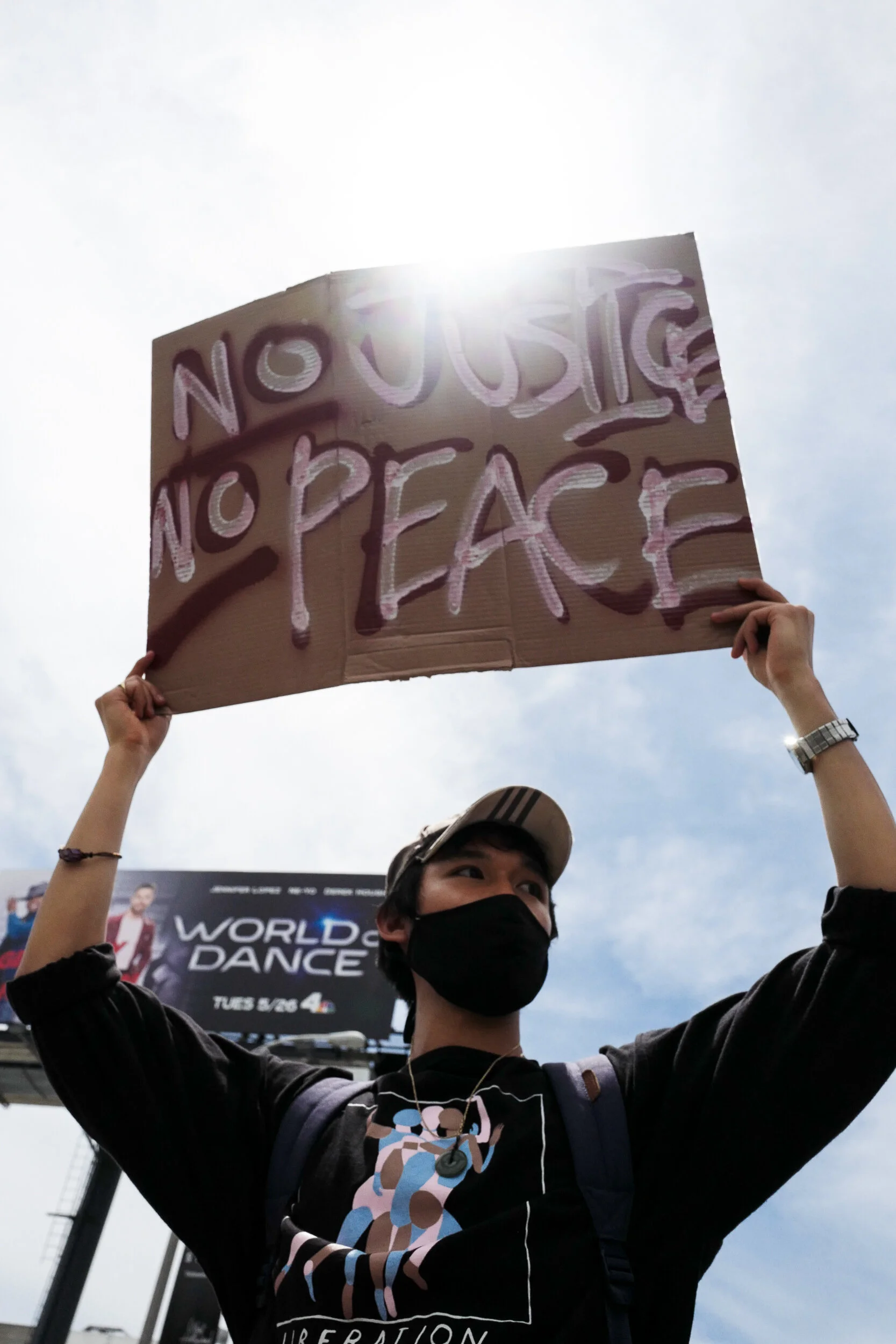 Person wearing a black face mask holding a cardboard protest sign that reads 'NO JUSTICE NO PEACE' during a demonstration, with a cloudy sky and a billboard in the background.