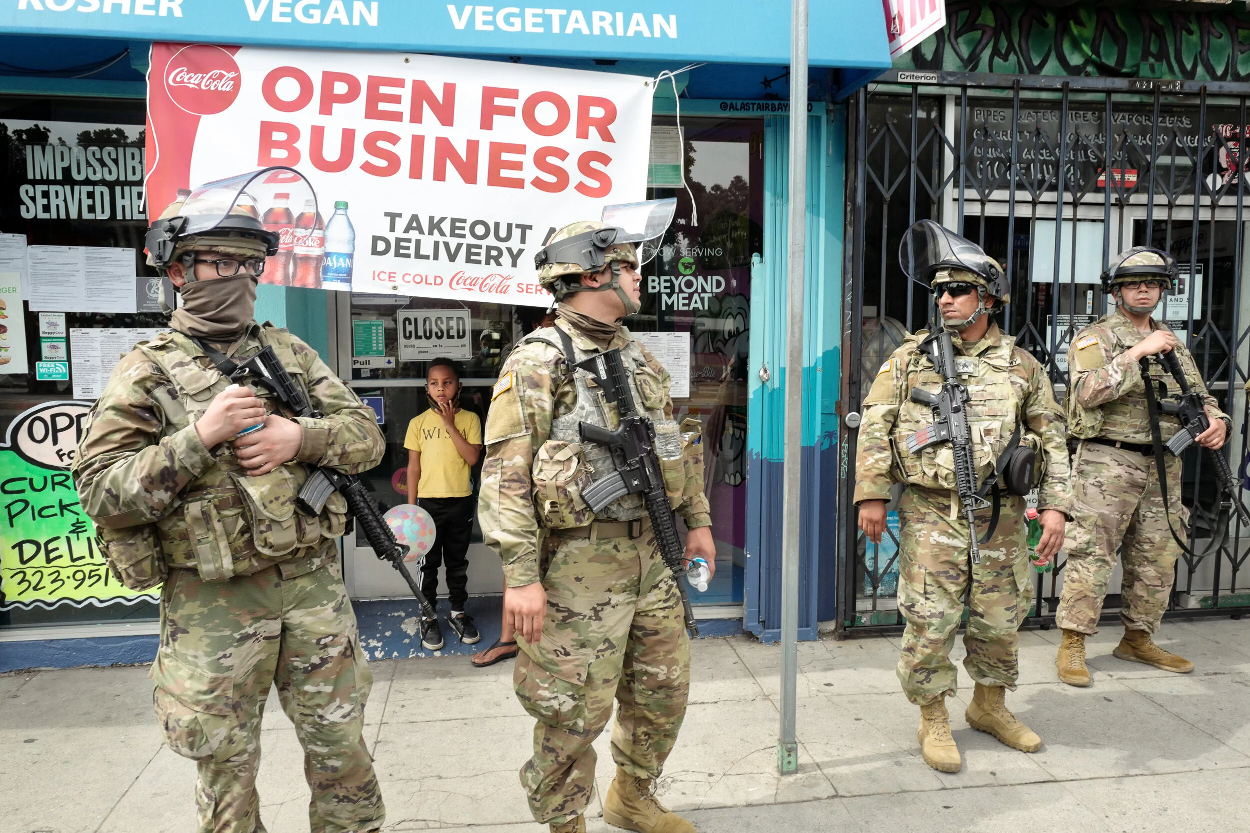 Four soldiers in camouflage uniforms and helmets standing guard on the sidewalk in front of a storefront with signs that read 'Open for Business' and 'Closed' in the window. Two children are visible behind the glass, one holding a balloon.