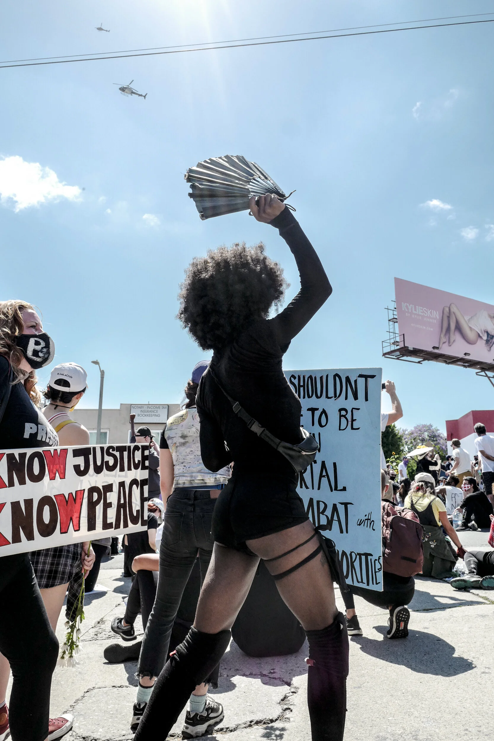 A woman with curly hair holds a fan up in the air at a protest, surrounded by other protesters holding signs with messages for justice and peace. The sky is clear with a helicopter flying overhead.