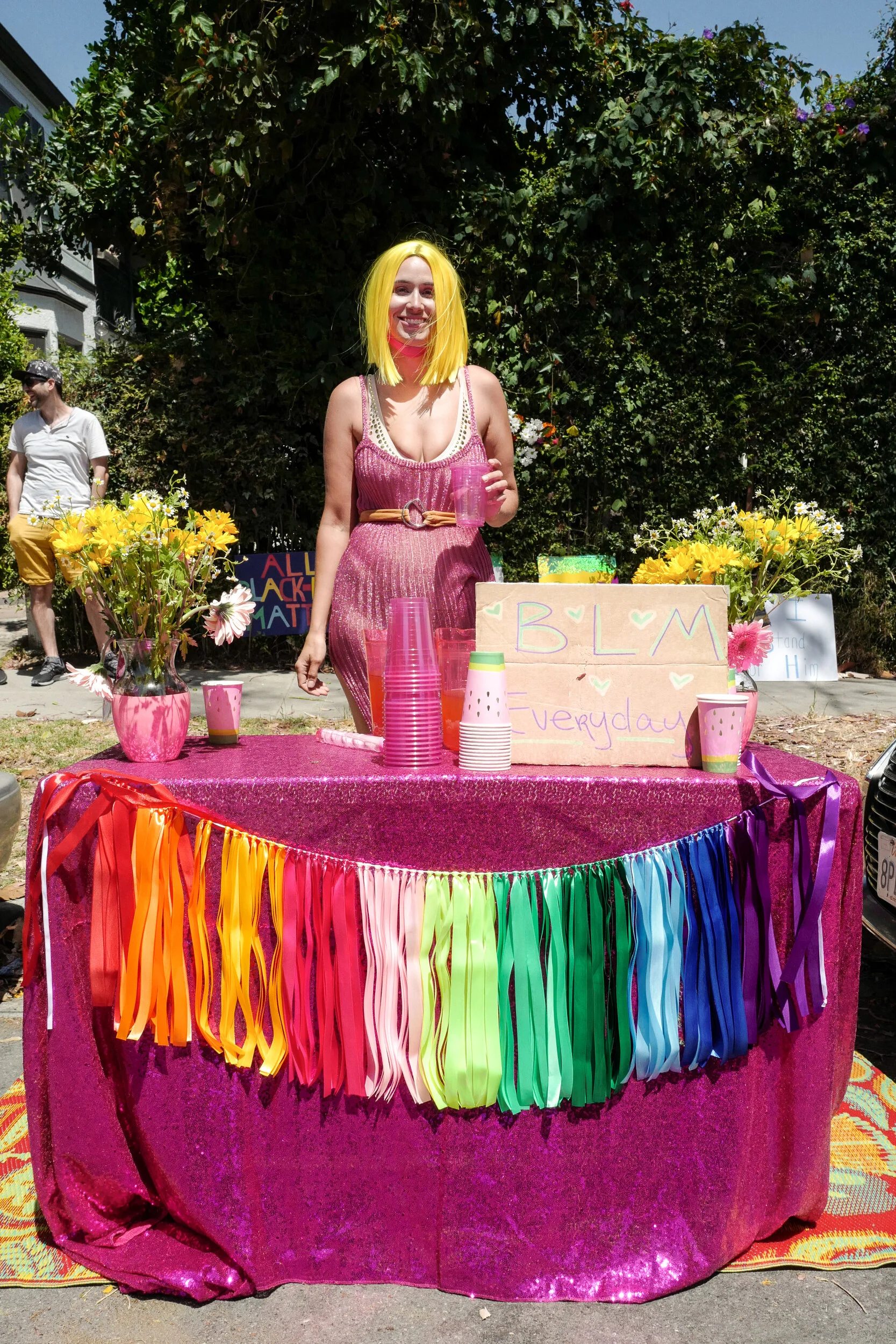 A woman with bright yellow hair wearing a pink dress standing behind a table decorated with pink, rainbow, and colorful streamers, with flowers and cups, at an outdoor event.