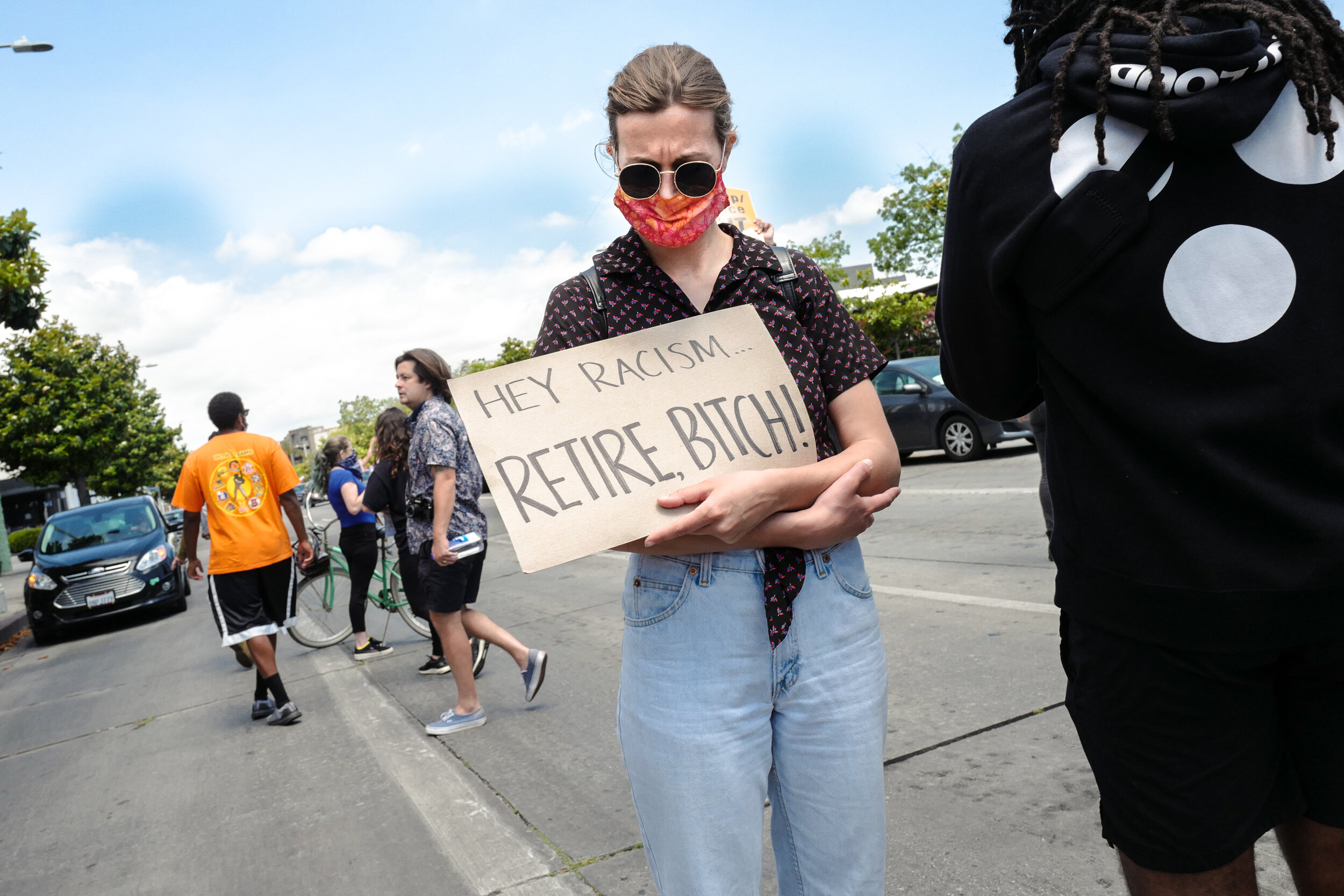 A woman wearing sunglasses and a face mask holding a cardboard sign that says, "Hey Racism... Retire, Bitch!" standing on a sidewalk during a protest or demonstration, with other people and cars visible in the background.