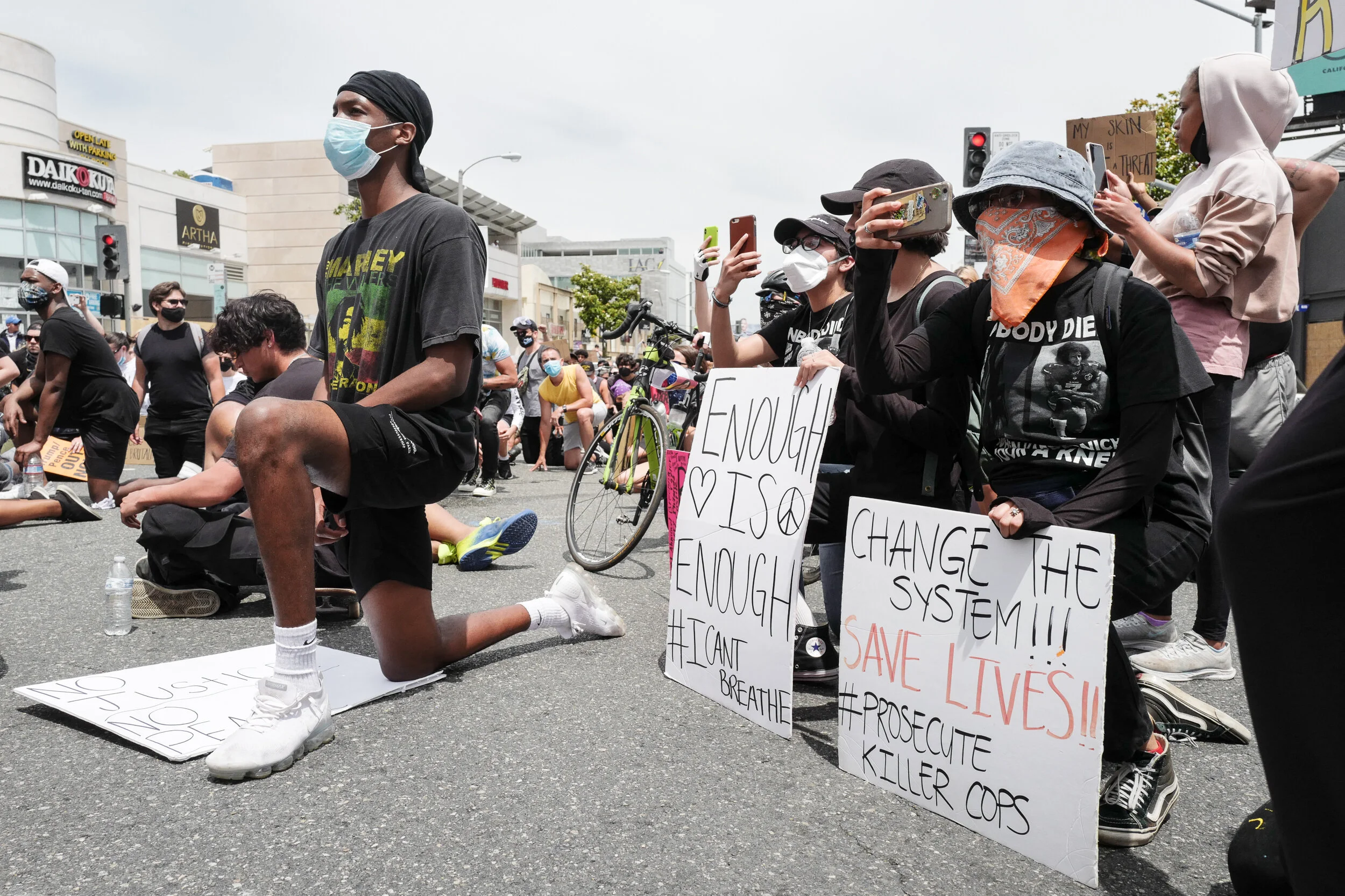Protesters kneeling and sitting in the street holding signs advocating for social justice and police reform, with some wearing masks and masks with messages, and others taking photos.