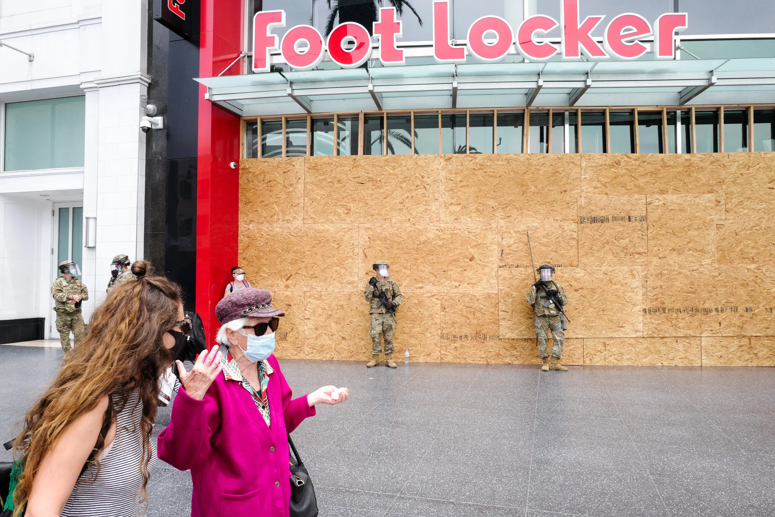 Foot Locker storefront with plywood boards covering the windows, armed military personnel standing guard, and a small crowd of people wearing masks and talking outside.