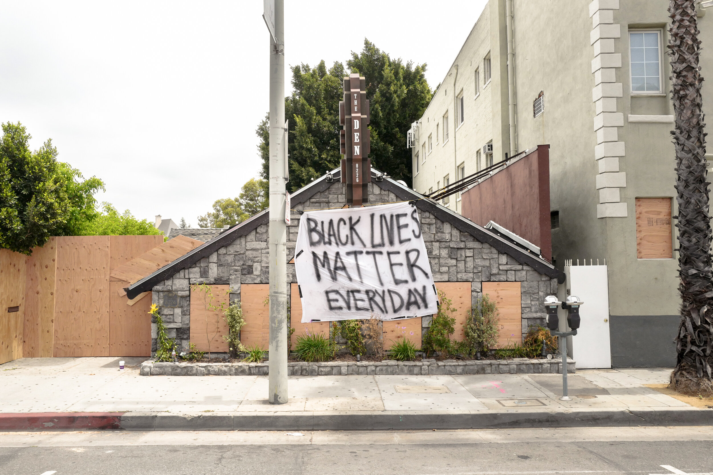 A house with a stone facade and boarded-up windows, with a large spray-painted banner that reads "Black Lives Matter Everyday!" hanging on the front. There are plants in front and a parking meter near the sidewalk.