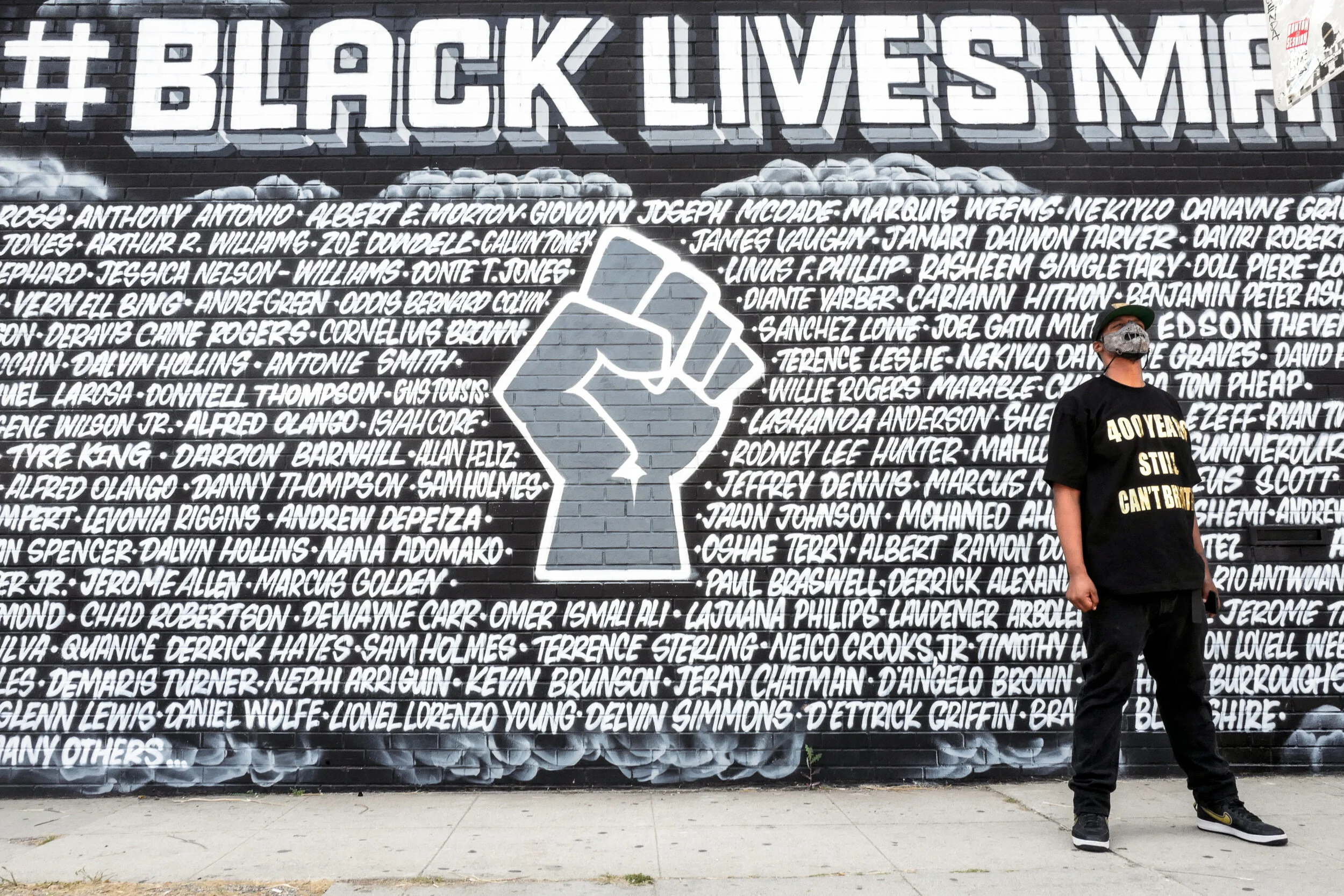 A man is standing in front of a large mural with hashtag #BlackLivesMatter and a white raised fist symbol on a black background, surrounded by numerous names of individuals.