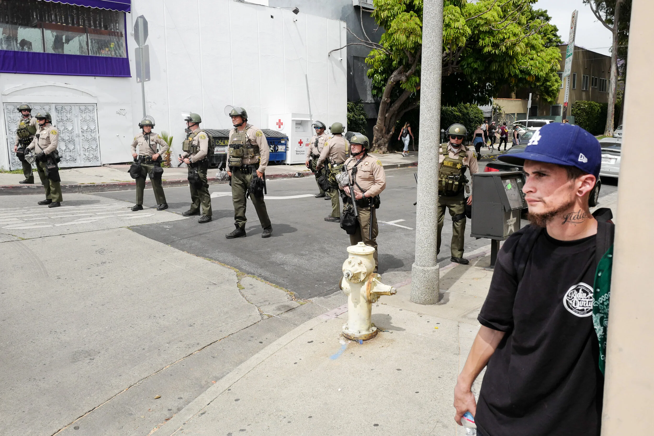 A man wearing a blue baseball cap and black shirt with a tattoo on his neck leans against a wall on the right side of the image while watching a row of police officers standing in the street. The officers are wearing tan uniforms, tactical gear, and 