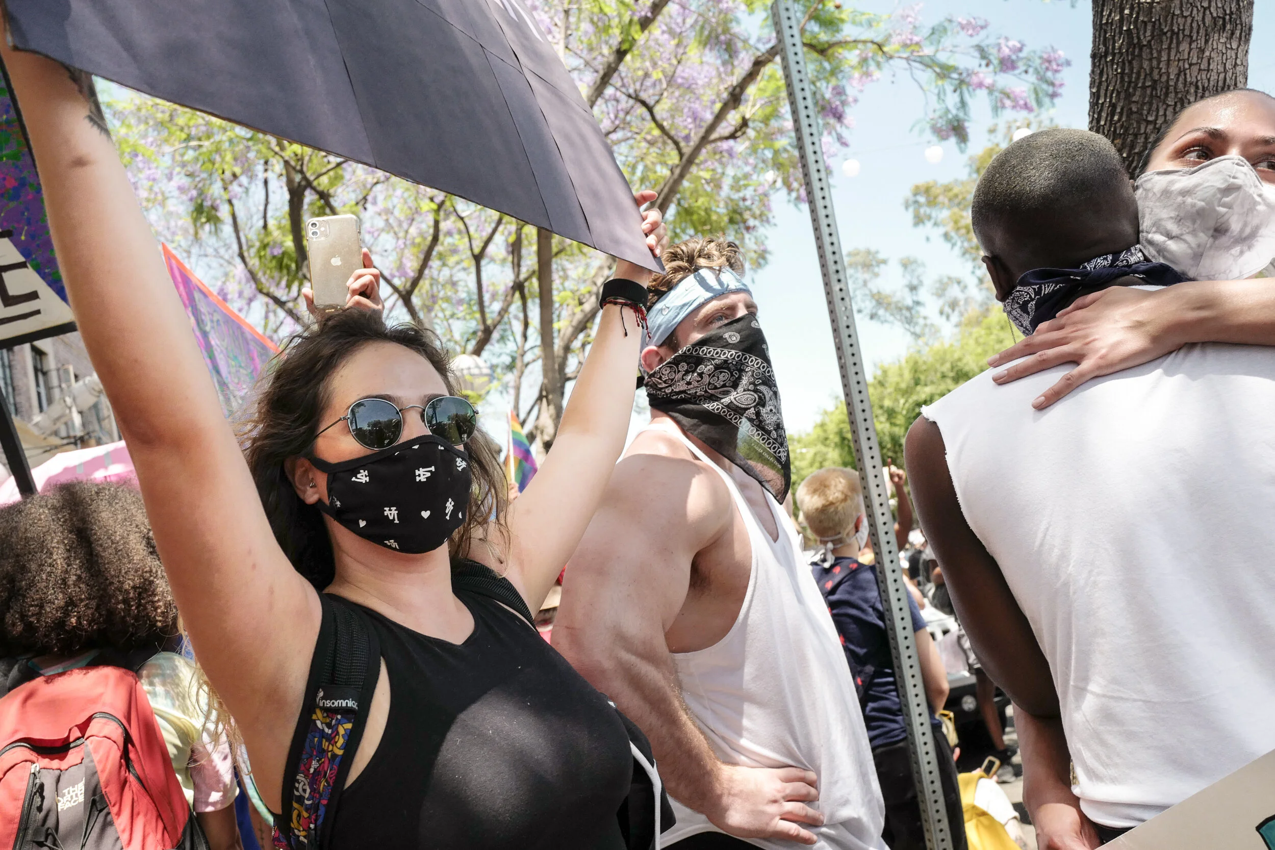 Multiple people at a protest or rally, some wearing face masks and bandanas, surrounded by trees and bright sky, with one woman holding a smartphone and holding an umbrella.
