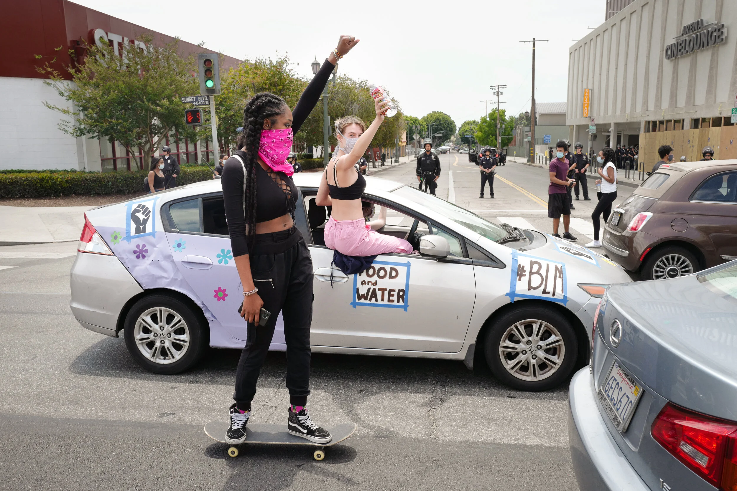 A protest scene with two young women in front of a silver car. One woman stands on a skateboard, wearing black clothes, a pink bandana as a face covering, and holding a smartphone. The other woman sits on the car's door frame, waving with her right a