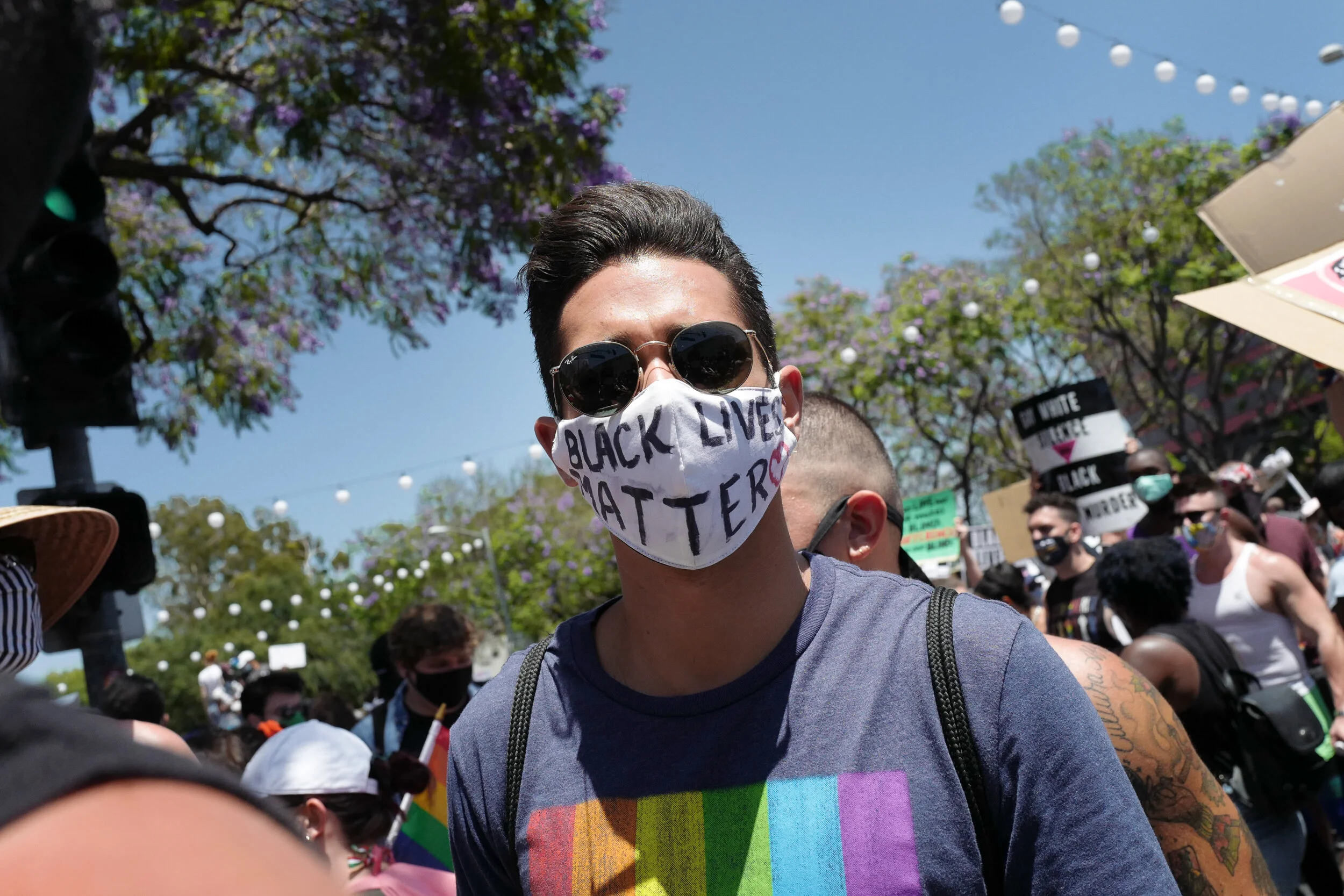 Person wearing sunglasses and a face mask with 'Black Lives Matter' written on it, participating in a protest or march with other demonstrators, some holding signs, outdoors on a sunny day with trees and string lights overhead.