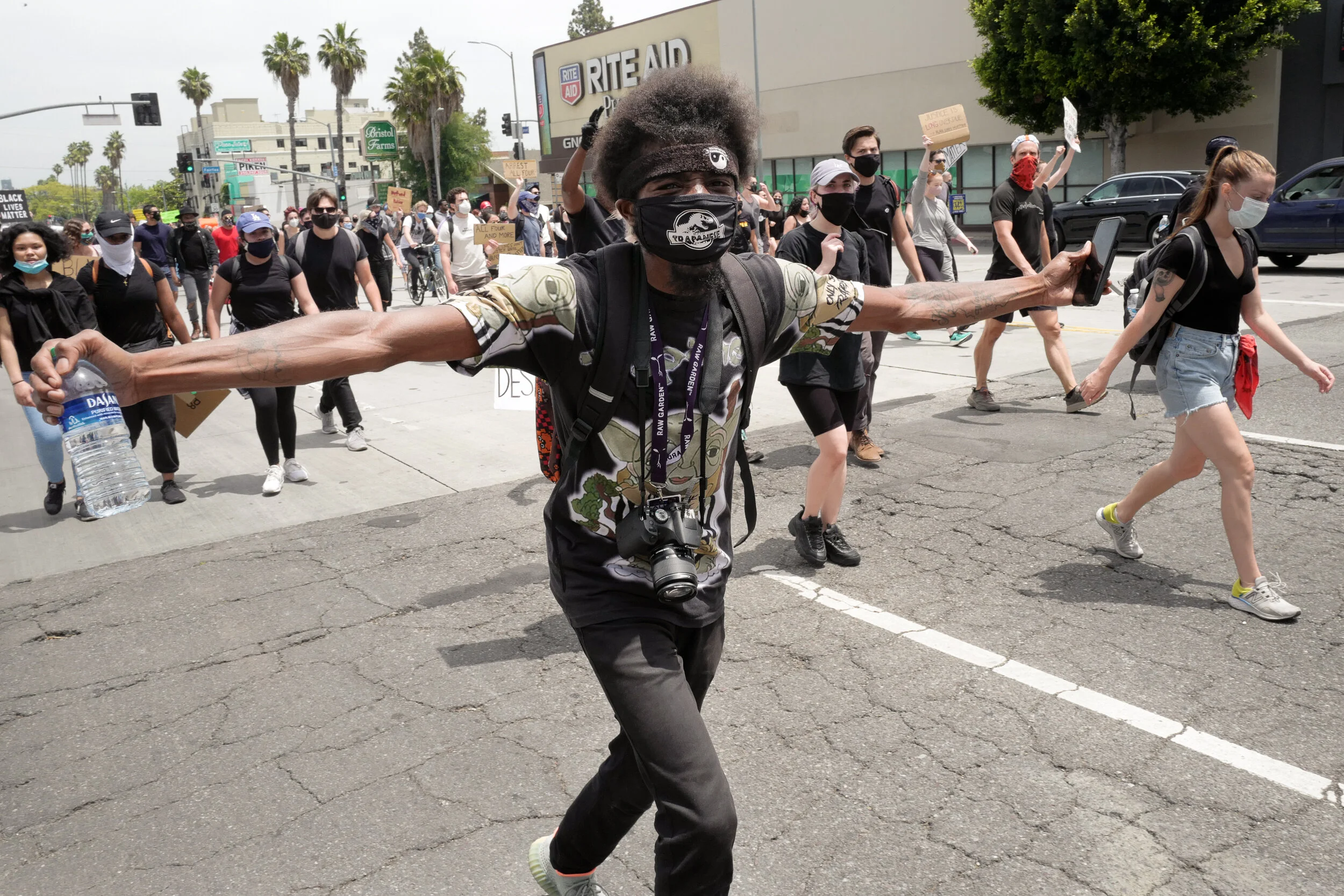 People participating in a protest march on the street, with some holding signs, wearing masks, and walking past a Rite Aid store.