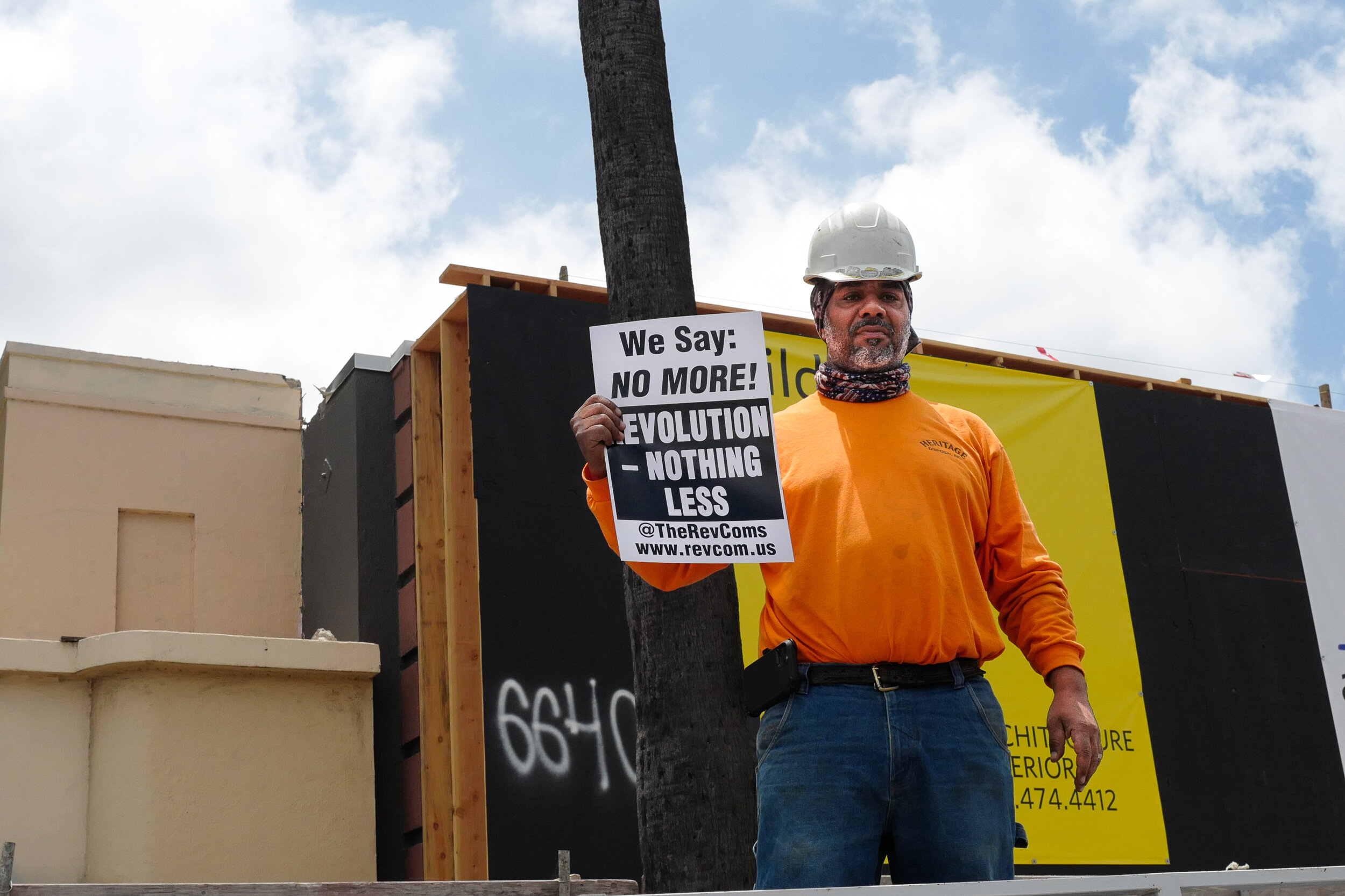 A man wearing a white hard hat, orange long-sleeve shirt, and jeans holds a protest sign that says 'We Say: NO MORE! EVOLUTION - NOTHING LESS' at a construction site with a partly cloudy sky in the background.