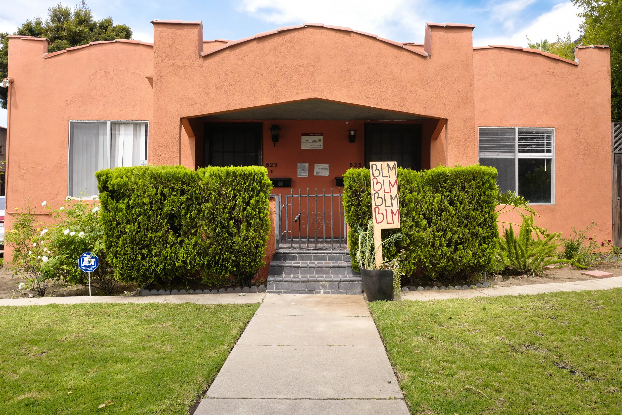 A pink stucco apartment building with a small front yard, green bushes, and a sidewalk. There is a protest sign with 