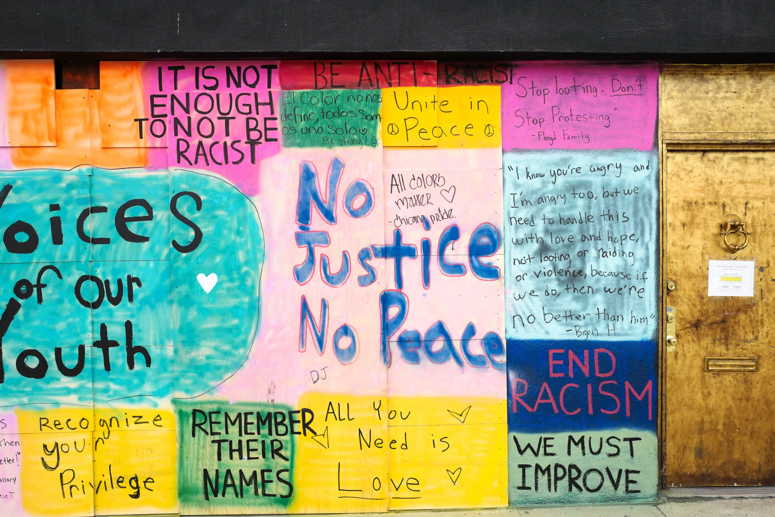 Colorful mural on a wall with messages promoting social justice, unity, peace, and anti-racism. Contains phrases like "No Justice No Peace," "End Racism," and "We Must Improve," along with handwritten notes and quotes.