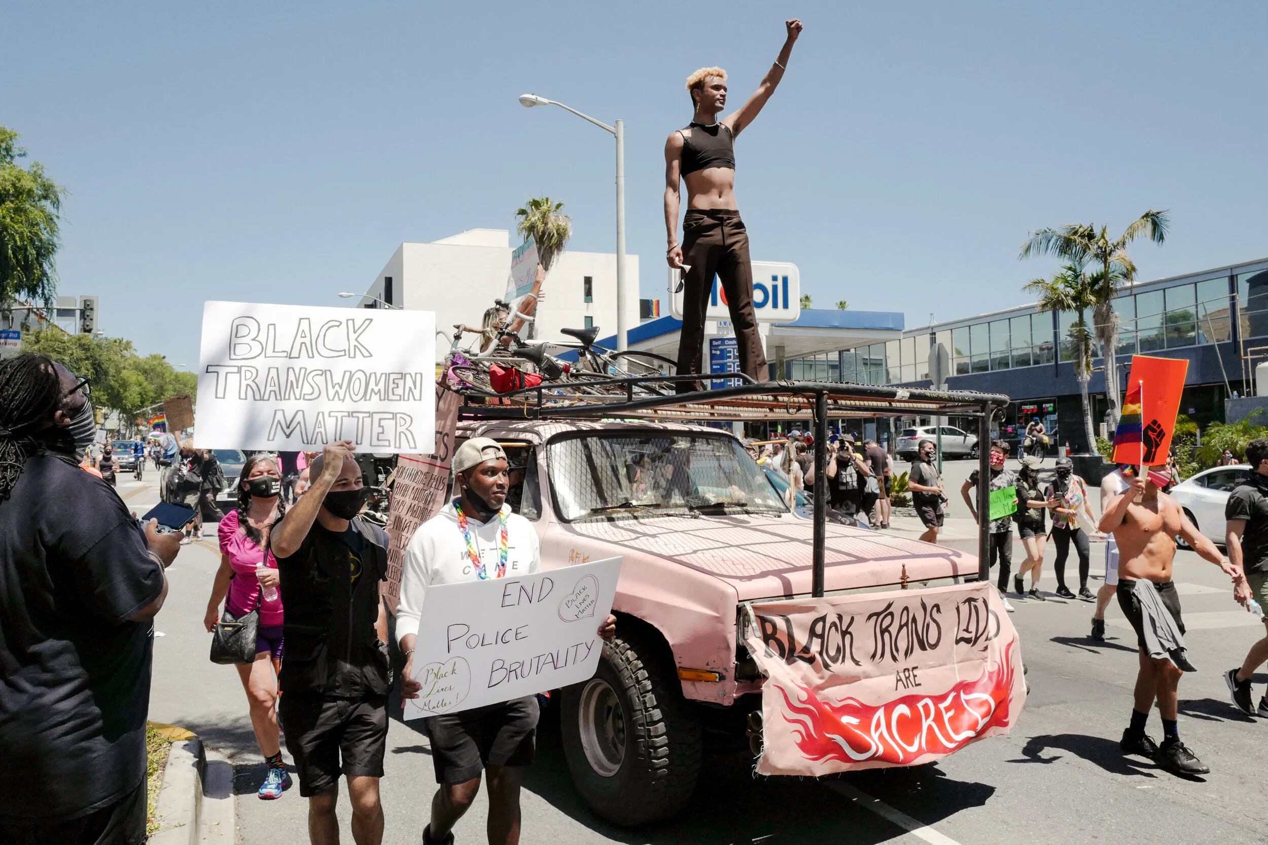 Protesters march during a demonstration for Black Trans Women Matter. They carry signs saying 'Black Trans Women Matter' and 'End Police Brutality.' A group of people, some shirtless, are walking alongside a pink vehicle adorned with signs and banner