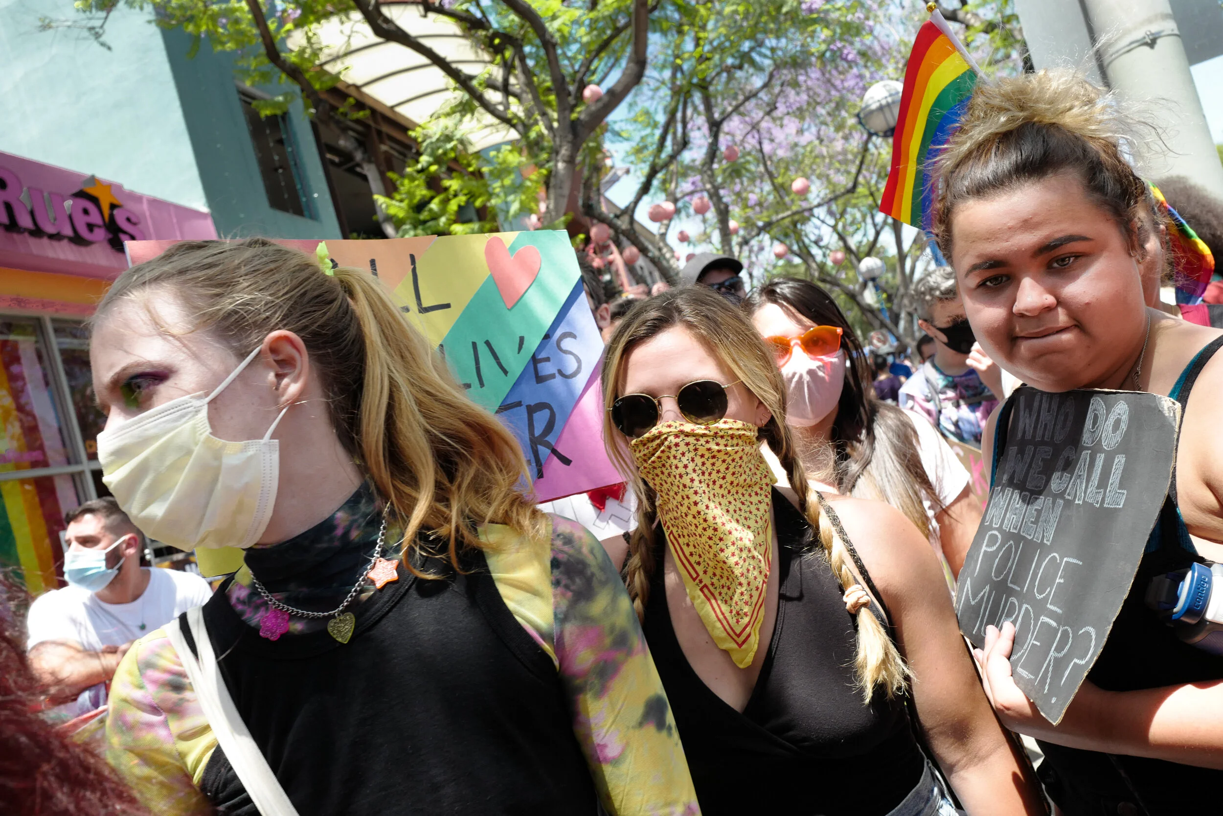 A diverse group of people at a pride parade, wearing masks and holding signs with messages supporting LGBTQ+ rights, with rainbow flags and colorful decorations in the background.