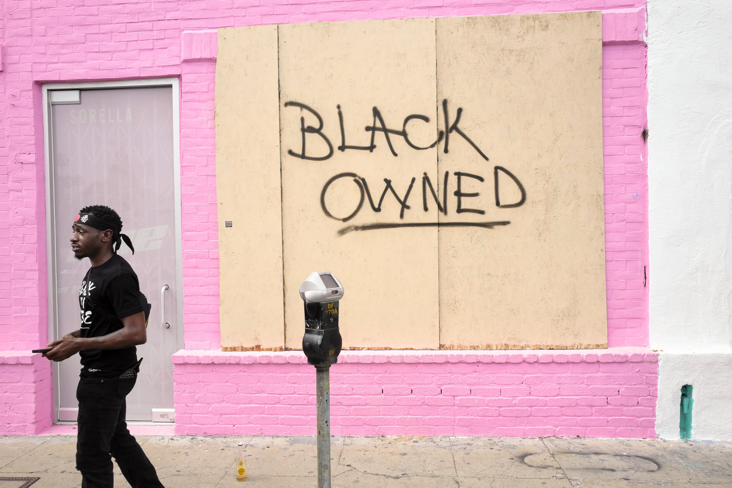 A man walking past a building with pink and white walls. The building has a boarded-up window and a graffiti message on the boarded-up section that reads 'Black Owned' in black spray paint.